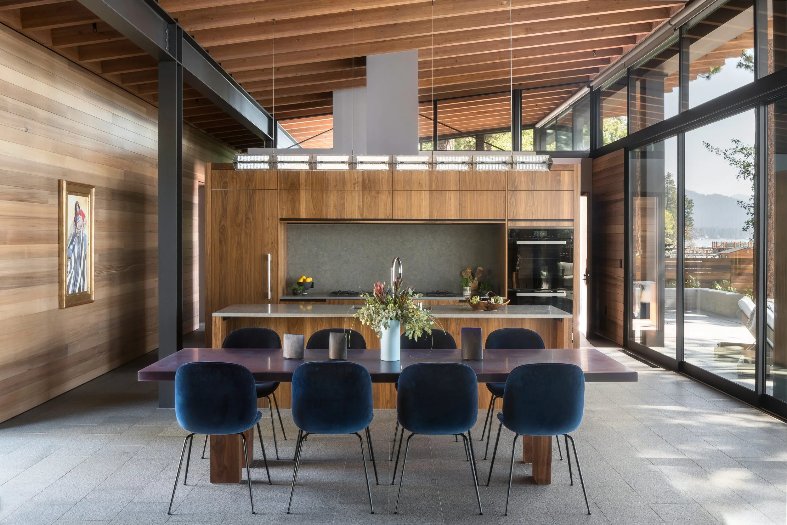 Modern dining area featuring a dark table with six blue velvet chairs, a large white vase with flowers, and a wooden kitchen backdrop with a gray backsplash and built-in oven. Large windows reveal an outdoor view of trees and water.