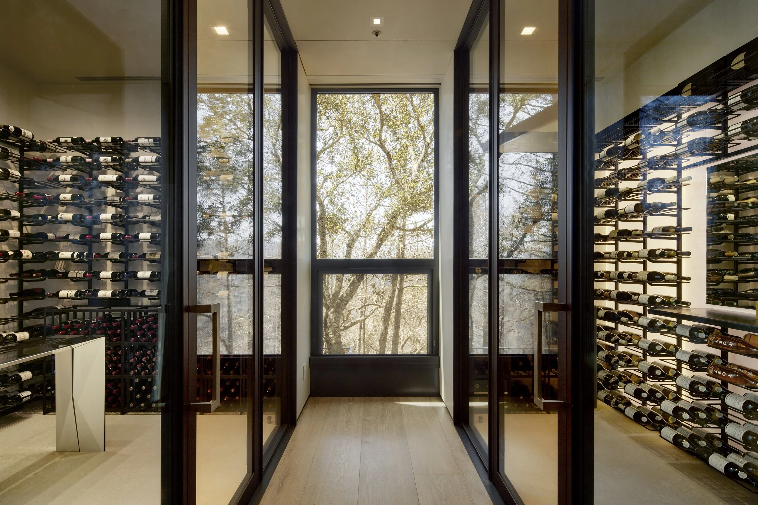 A wine cellar with floor-to-ceiling wine racks on both sides, glass doors at the end revealing trees outside, and natural light coming through the windows.