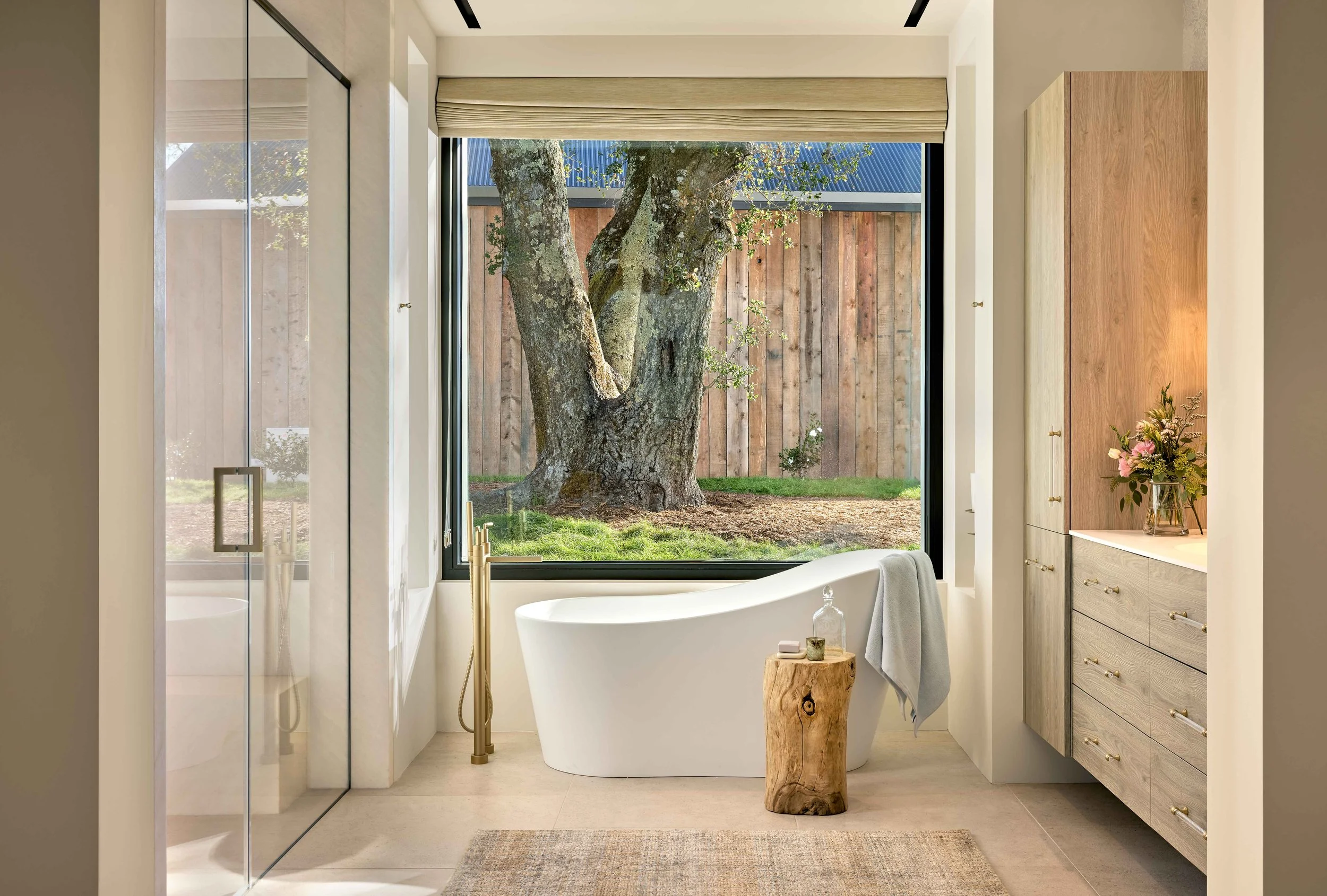 Bathroom with a view of a large tree outside the window, featuring a bathtub, wooden cabinet, flower arrangement, and a small wooden stool.