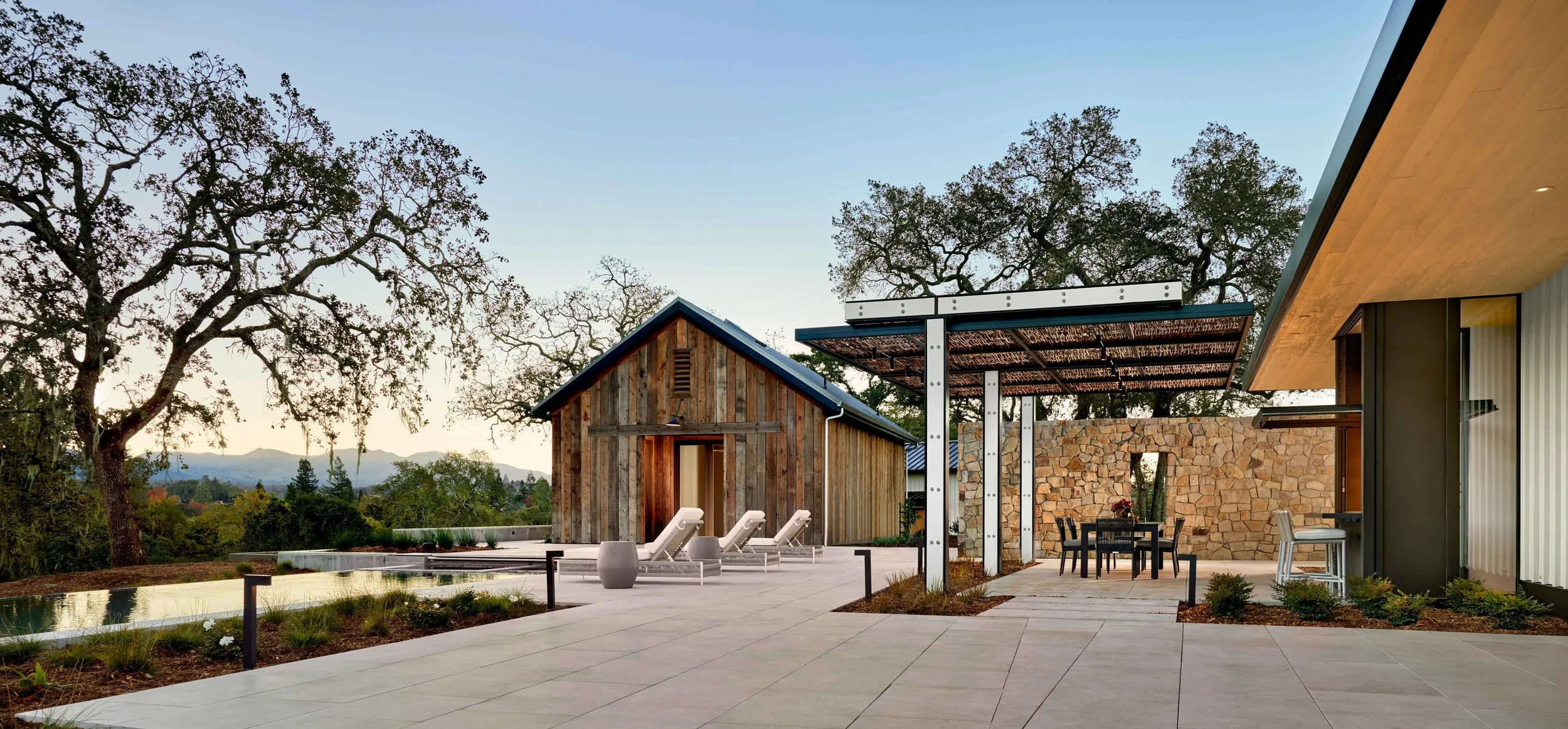 Modern residential outdoor space with a pool, patio, lounge chairs, dining area, wooden shed, stone wall, and trees in the background during sunset.