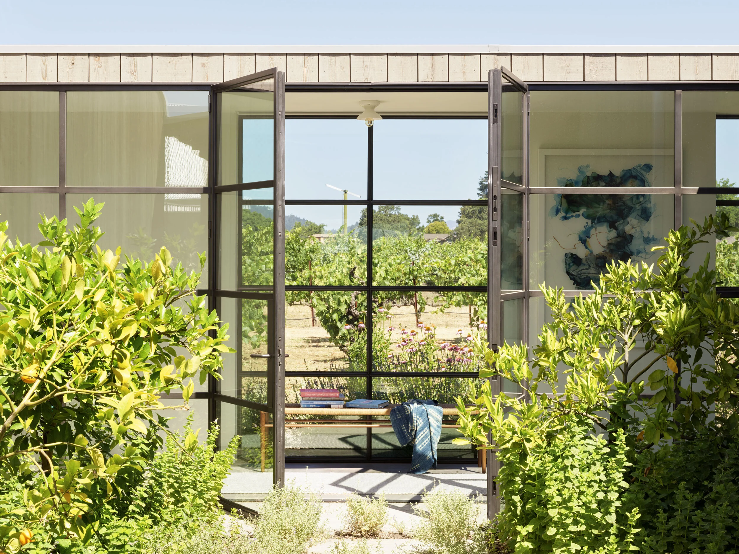Open glass door leading to a garden with lush greenery and flowering plants, with a house and blue sky in the background.
