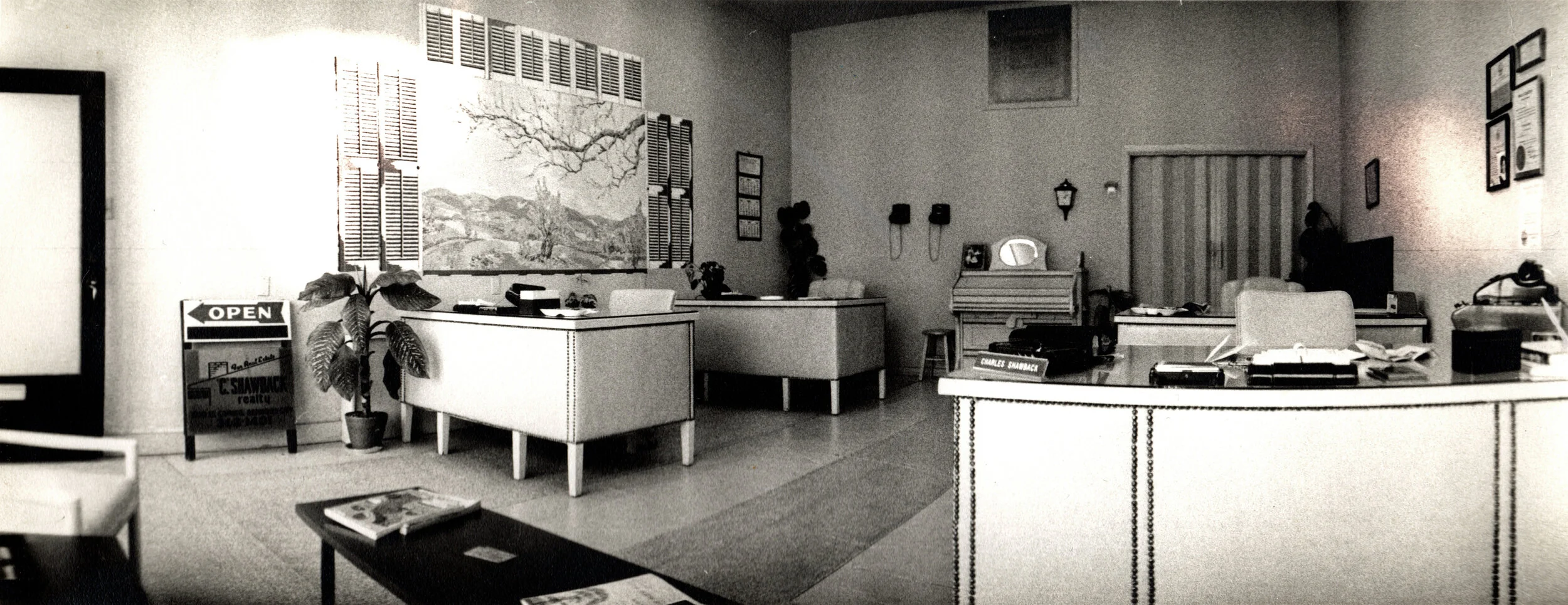 An office reception area with a desk, chairs, artwork on the wall, a potted plant, and various office supplies.