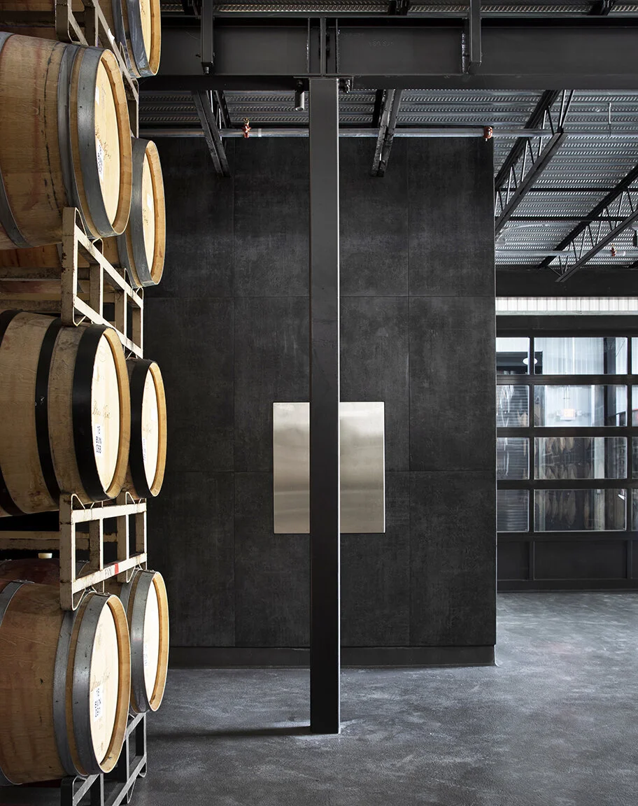 Empty wine cellar or storage room with stacked wooden barrels on the left and a large black wall with a metallic plaque in the center. There are windows on the right side allowing natural light into the space.