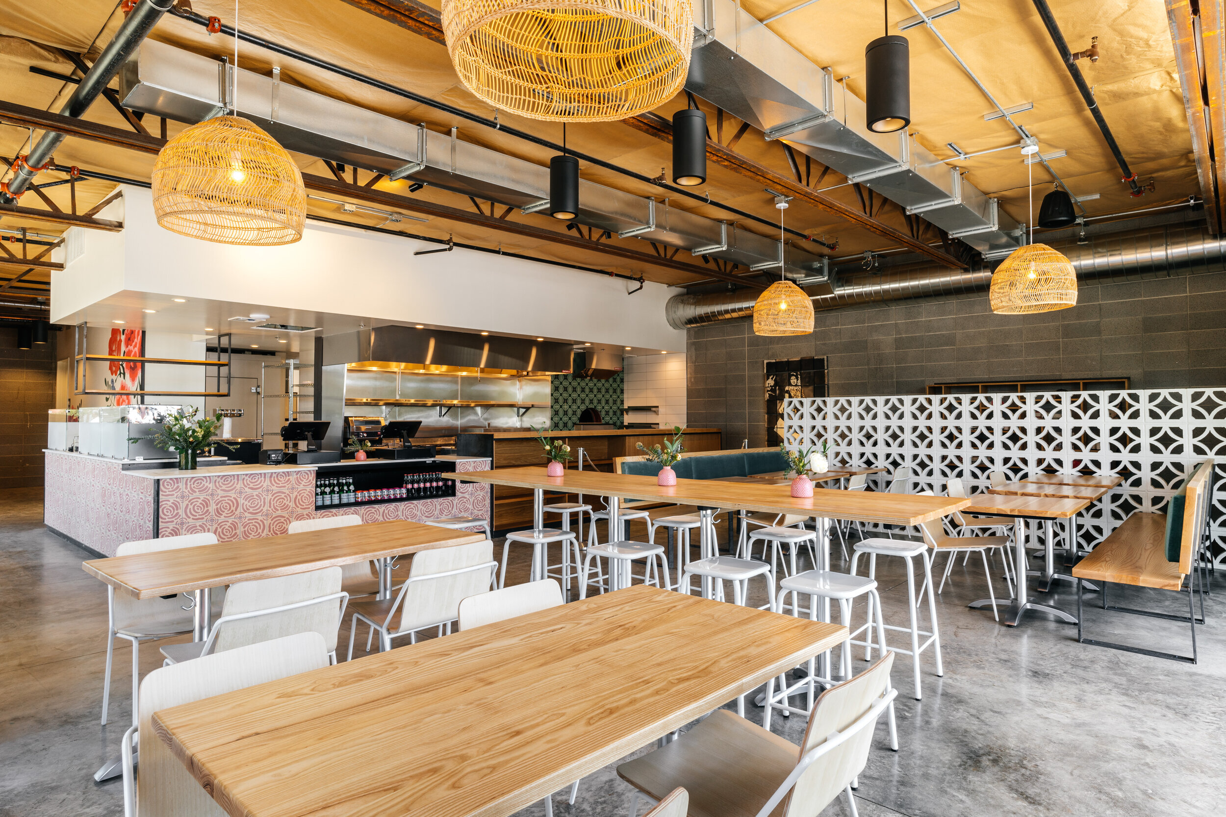 Inside view of a modern restaurant with wooden tables, white chairs, hanging wicker lights, and an open kitchen area with stainless steel surfaces and green tiles.