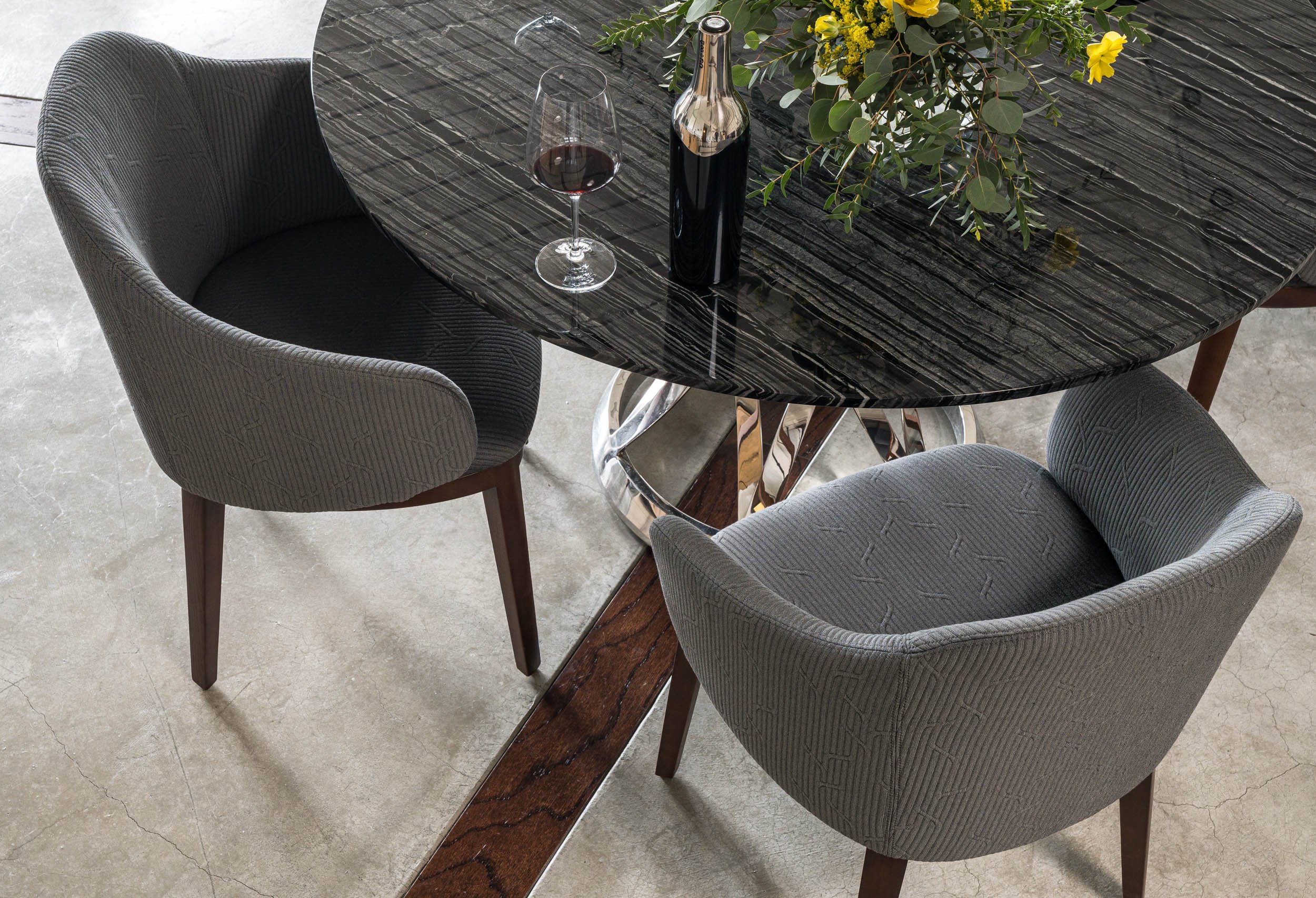 A dining table with a black marble top, surrounded by four gray upholstered chairs. On the table, there is a glass of red wine, a wine bottle, and a floral arrangement with yellow flowers and greenery. The table has a chrome base, and the floor is light-colored concrete.