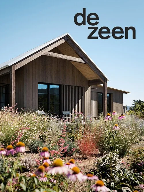 A modern wooden house with a sloped roof, large windows, and a porch, surrounded by colorful wildflowers in a rural setting under a clear blue sky.