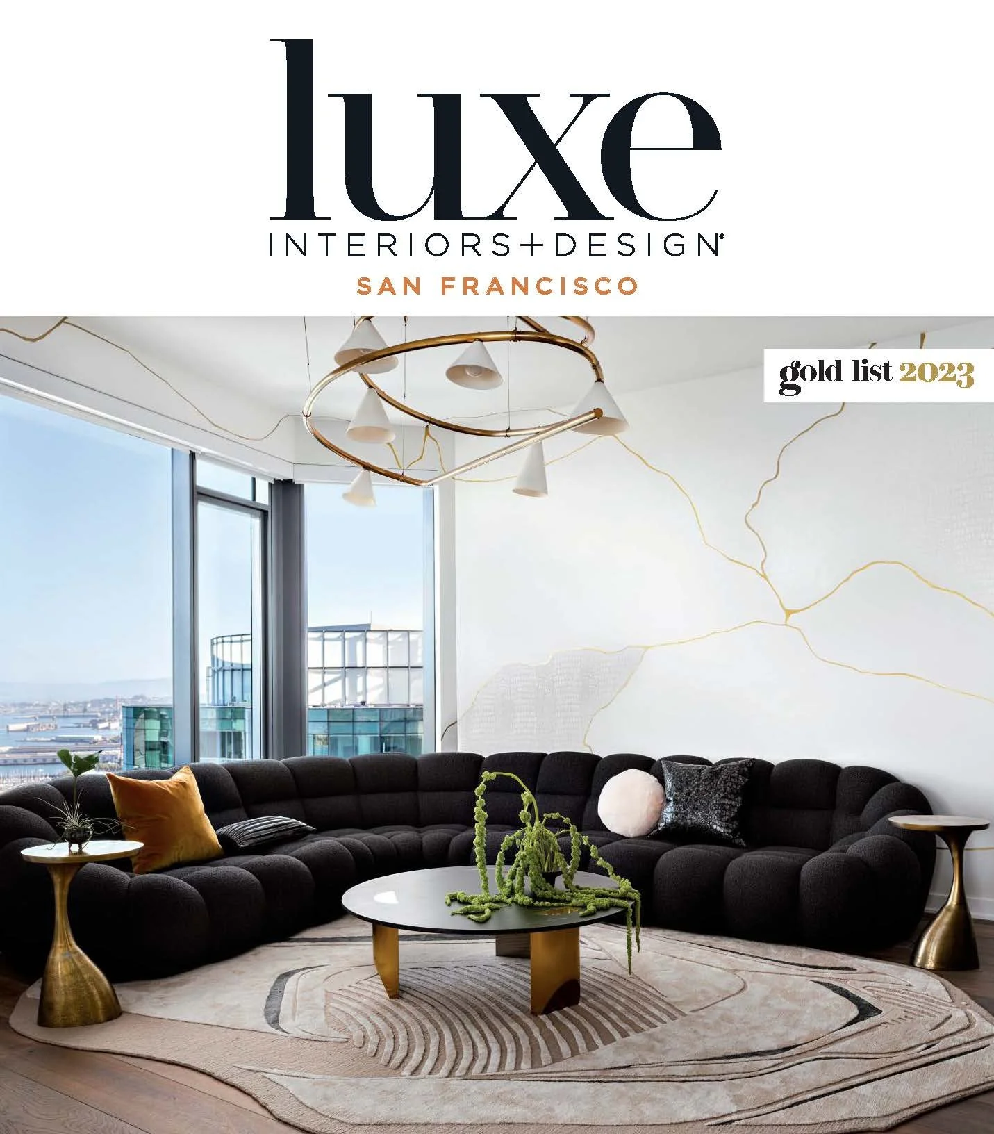 Interior living room with large black sectional sofa, gold side tables, a white coffee table with a green plant sculpture, pendant lighting, and large windows showing a city view.