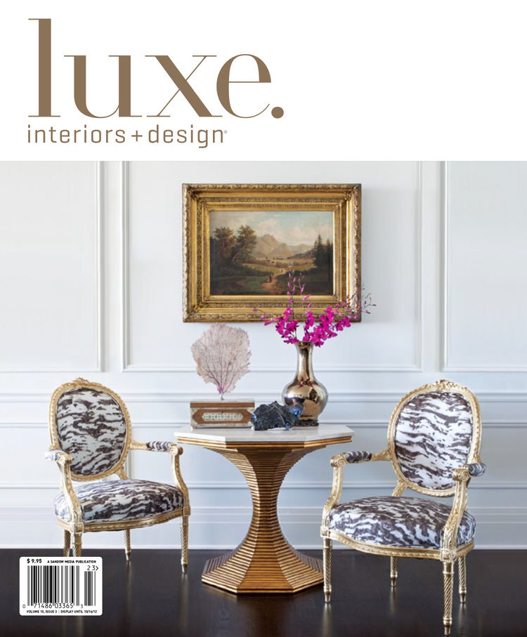 Cover of Luxe Interiors + Design magazine featuring a classic interior scene with a round wooden table, two ornate chairs with zebra print upholstery, a large gold vase with pink flowers, a small pink coral sculpture, and a landscape painting in an o