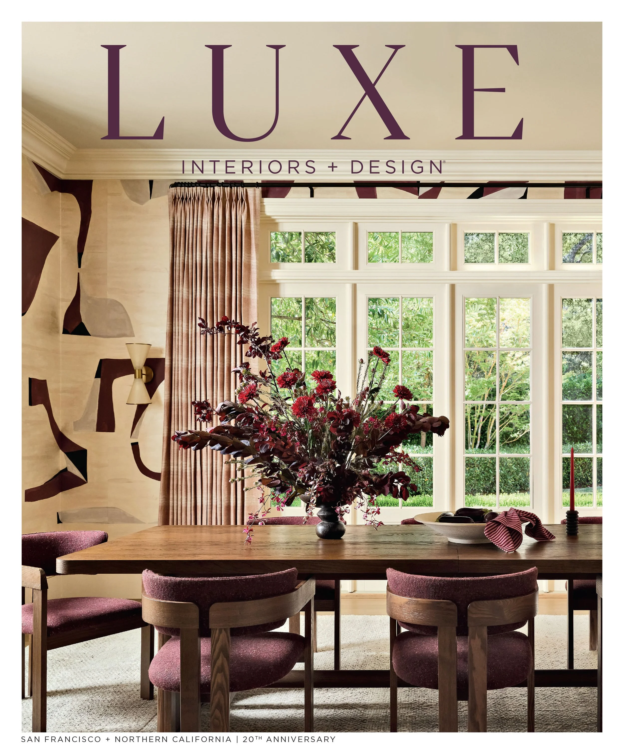 A dining room with a wooden table and six chairs with purple upholstered seats and backrests. On the table, there is a floral arrangement, a bowl of fruit, and a tall candle in a holder. Behind the table, there are large windows with a view of green 