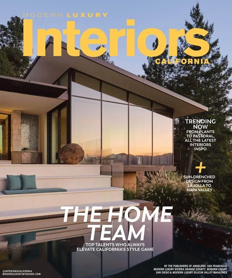 Modern luxury home with large windows reflecting a sunset, surrounded by greenery and outdoor seating, on the cover of California interior design magazine.