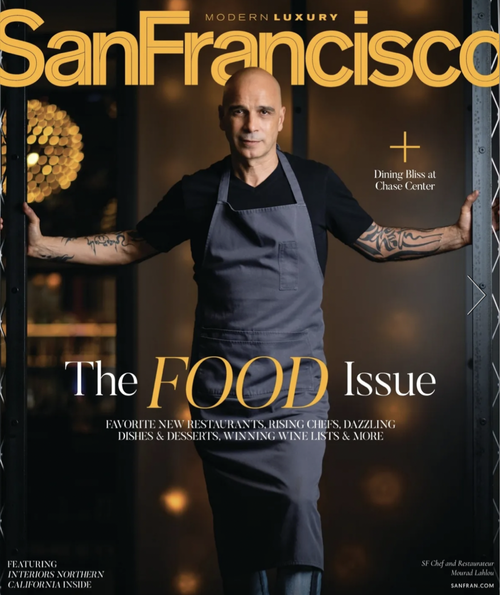 A man standing with arms outstretched, wearing a black shirt and gray apron, in front of a magazine cover titled San Francisco with the headline 'The Food Issue'.