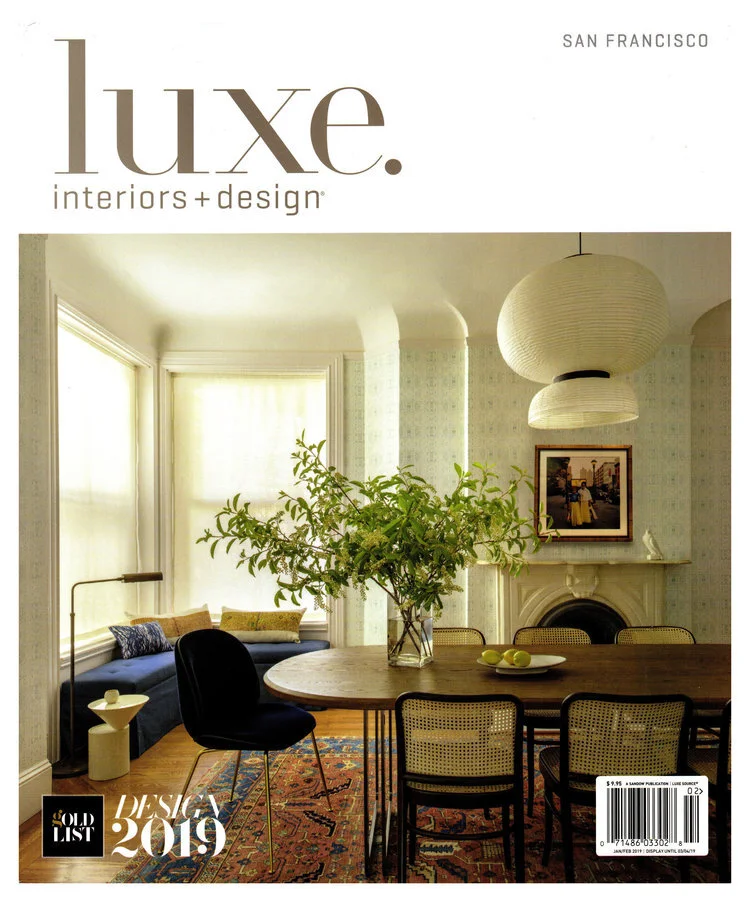 Cover of Luxe Interiors + Design magazine featuring a cozy dining area with a large wooden table, a glass vase with green foliage, a pale yellow or cream-colored paper lantern pendant, a fireplace, framed artwork, and a seating nook with cushions.
