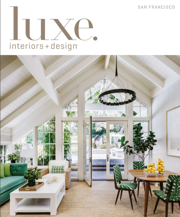 Interior view of a bright, airy sunroom with white vaulted ceiling, large windows, and glass door leading outside. The room features a green sofa, white coffee table, wicker armchair, dining table with green and white leaf-patterned chairs, and decor