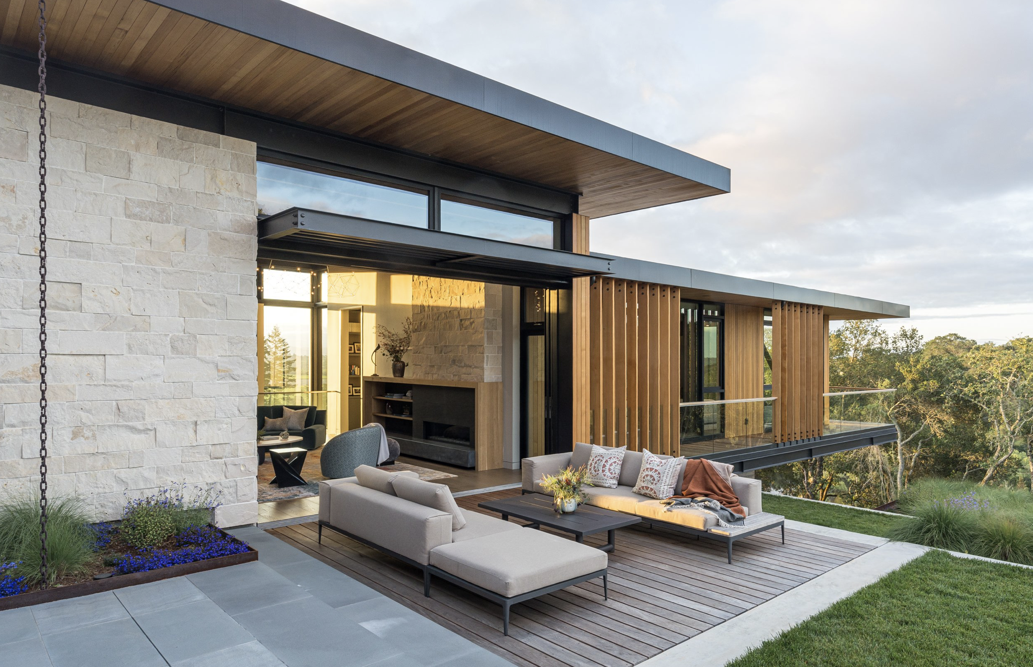 Modern house with outdoor patio, beige stone wall, glass sliding doors, wooden slat accents, and outdoor living furniture including a sofa, armchair, and coffee table, with trees and blue sky in the background.