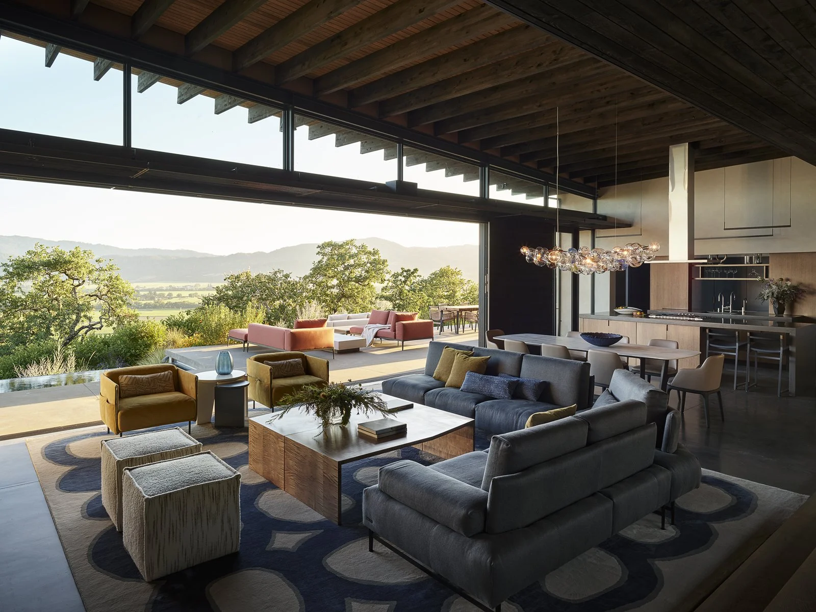 Modern living room with large glass doors opening to a scenic outdoor patio overlooking mountains and trees, featuring sofas, armchairs, a coffee table, and a contemporary chandelier.