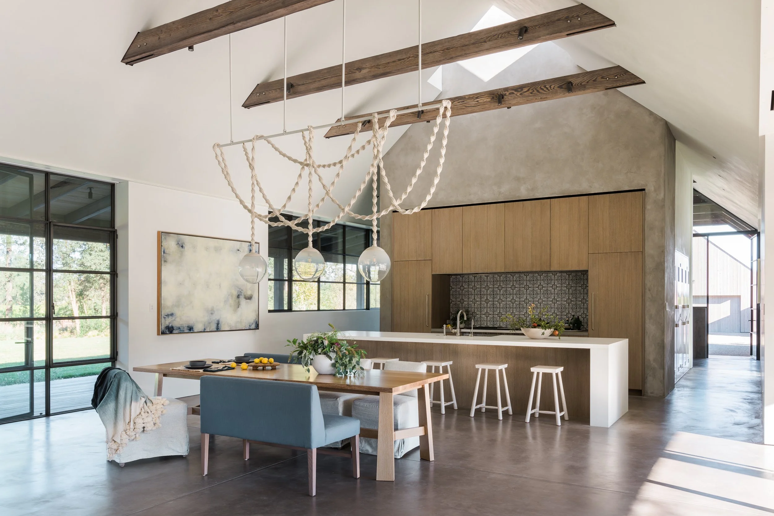 Modern open-concept kitchen and dining area with large windows, wooden beams, a kitchen island, and contemporary furniture.
