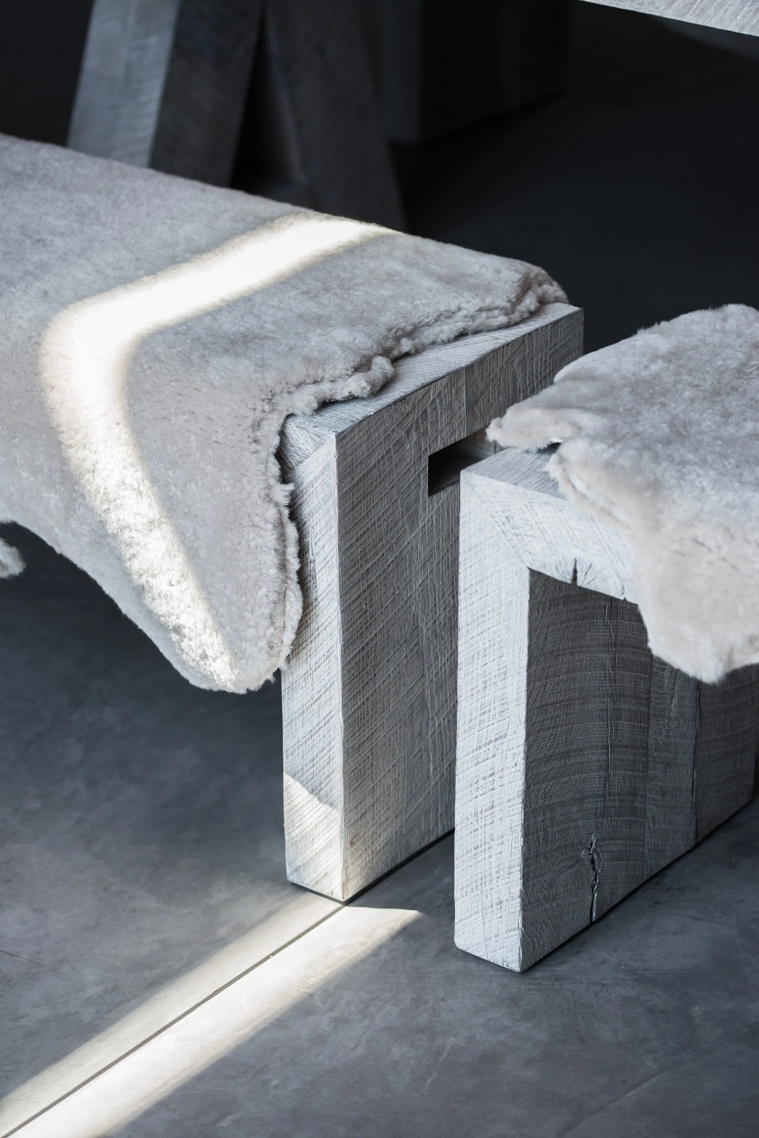 Two wooden benches with light-colored, plush blankets draped over them, set against a dark background and illuminated by a shaft of natural light.