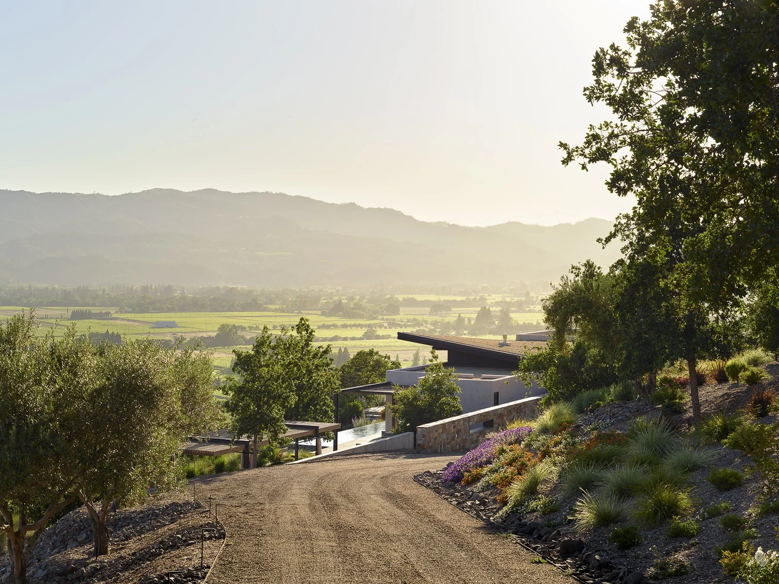 A scenic view of a modern house with a sloped roof surrounded by a garden of flowers and trees, with rolling hills and mountains in the background during sunset.