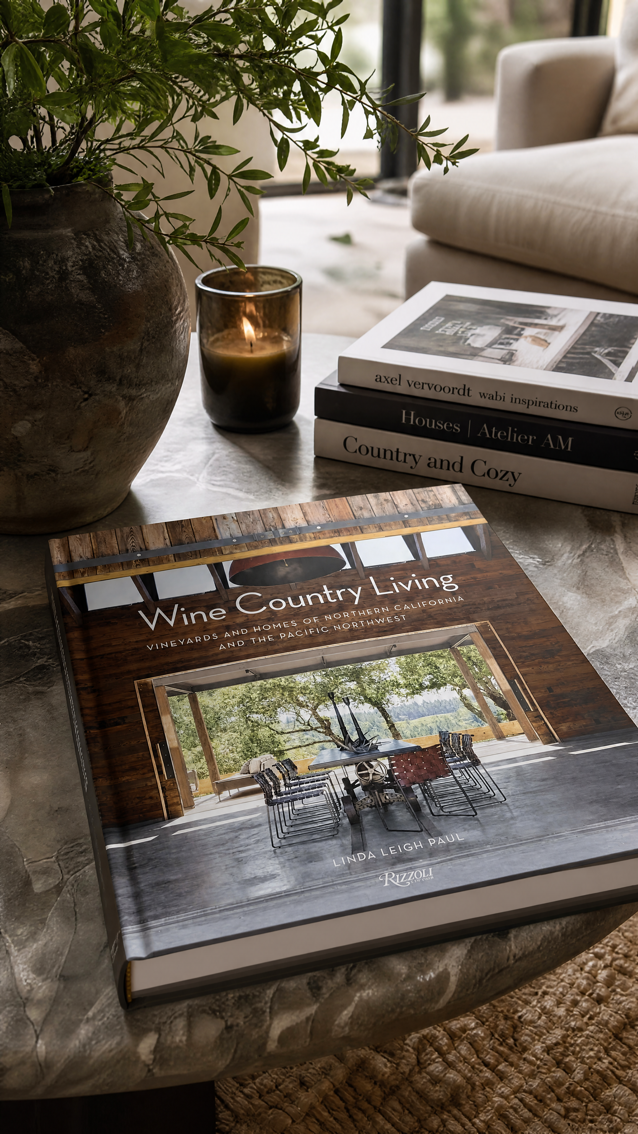 Coffee table with a green plant in a large rustic vase, a lit candle in a glass holder, and a stack of three books including one titled 'Wine Country Living' and a large magazine or book with a photo of an interior space on top.