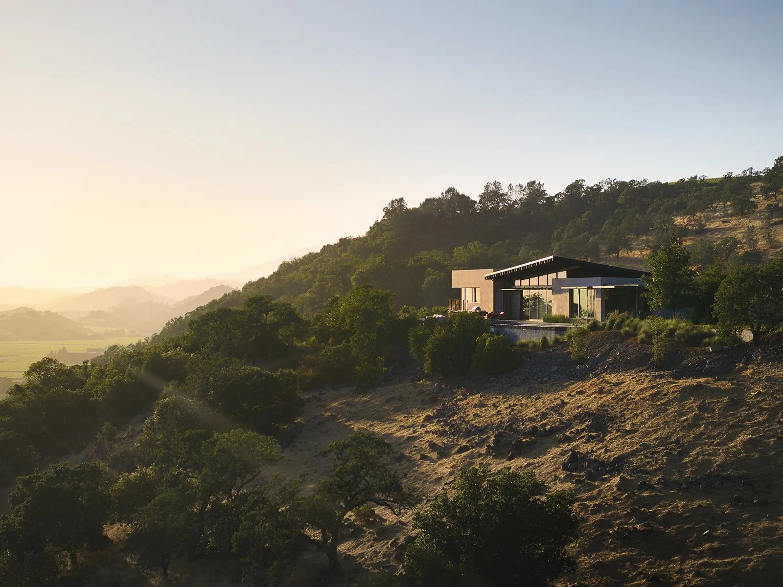 Modern house on a hillside surrounded by trees with a mountain range in the background during sunset.