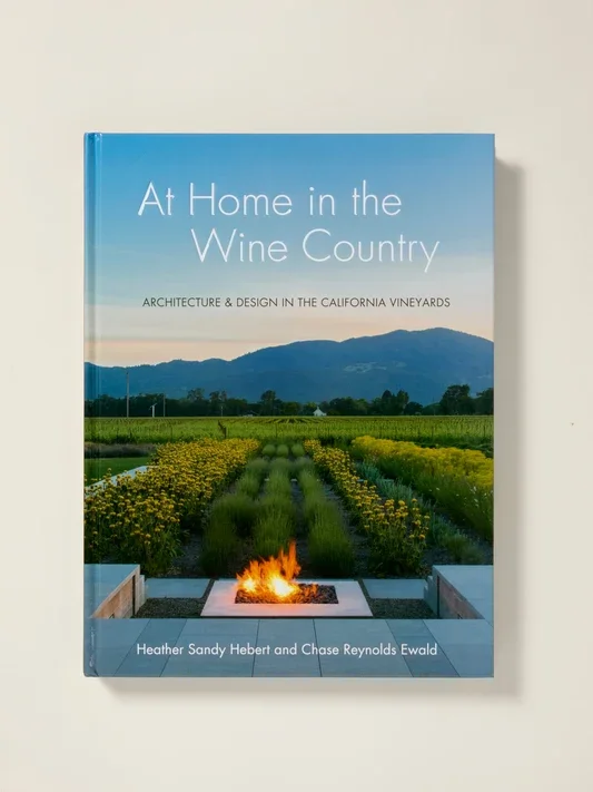 Book titled 'At Home in the Wine Country' featuring a vineyard landscape with mountains in the background, a fire pit in the foreground, and a clear sky.