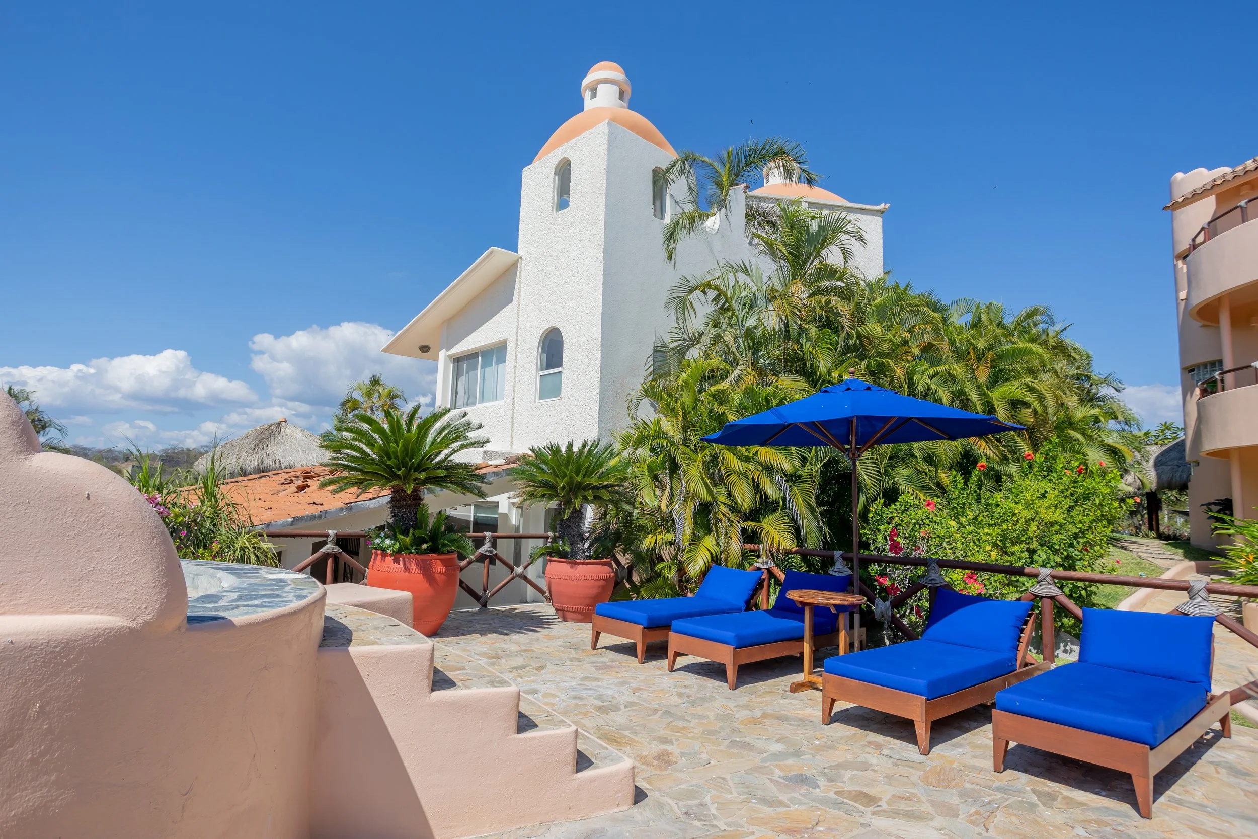 Outdoor patio with blue cushioned chairs, a blue umbrella, lush green plants, a white building with a dome, and a clear blue sky.