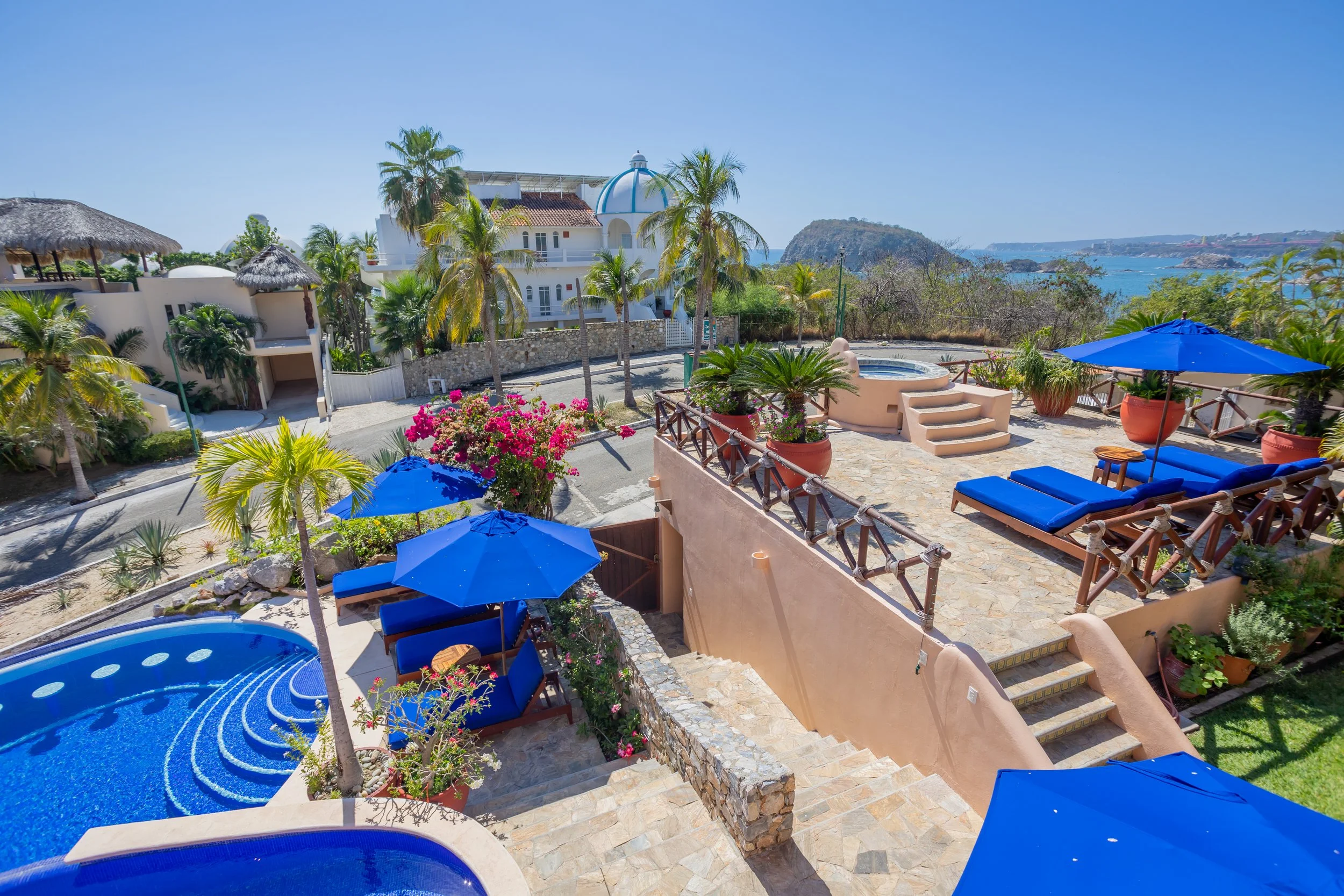 View of a sunny patio with blue lounge chairs, umbrellas, potted plants, and steps leading down to a small pool with a decorative blue design, overlooking a coastal landscape with water, hills, and a clear blue sky.
