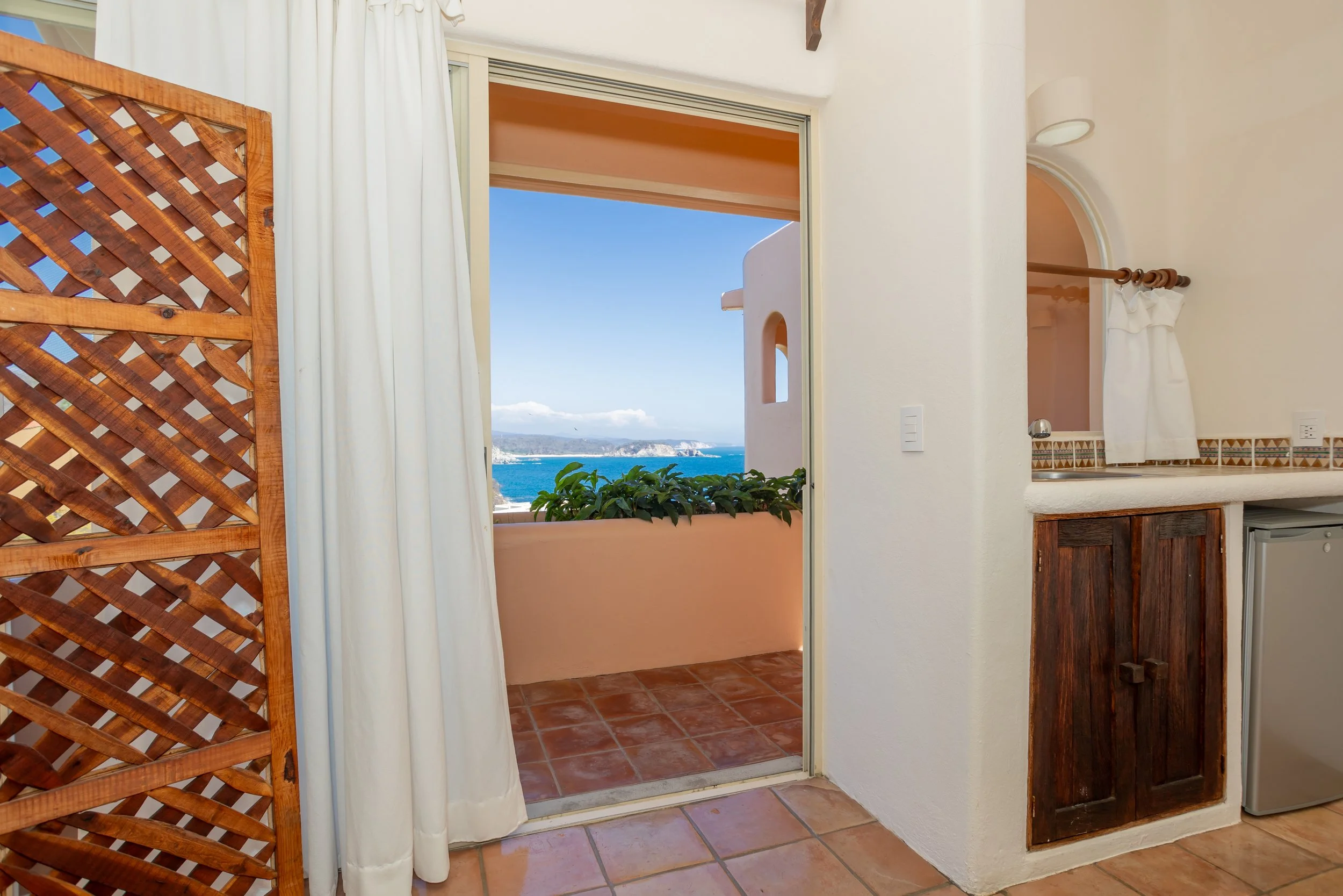 View through an open door to a balcony with a blue ocean and sky in the distance. The balcony has a plant, and the room has terracotta tile flooring, white walls, a white curtain, and wooden accents.