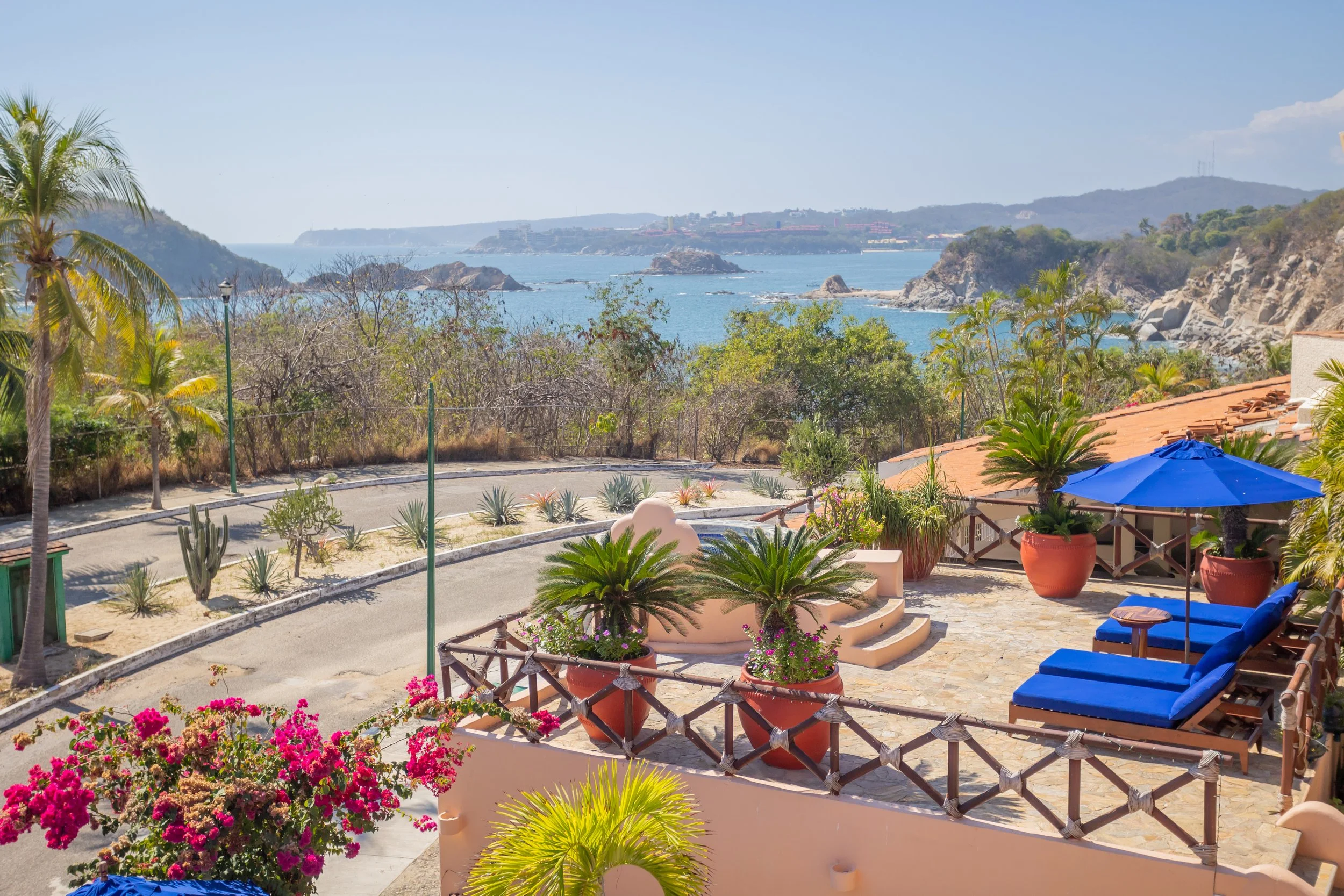 A scenic view of a coastal landscape with rocky cliffs, blue ocean, and a clear sky in the background, viewed from a patio area decorated with colorful flowering plants, green palm trees, and blue lounge chairs.
