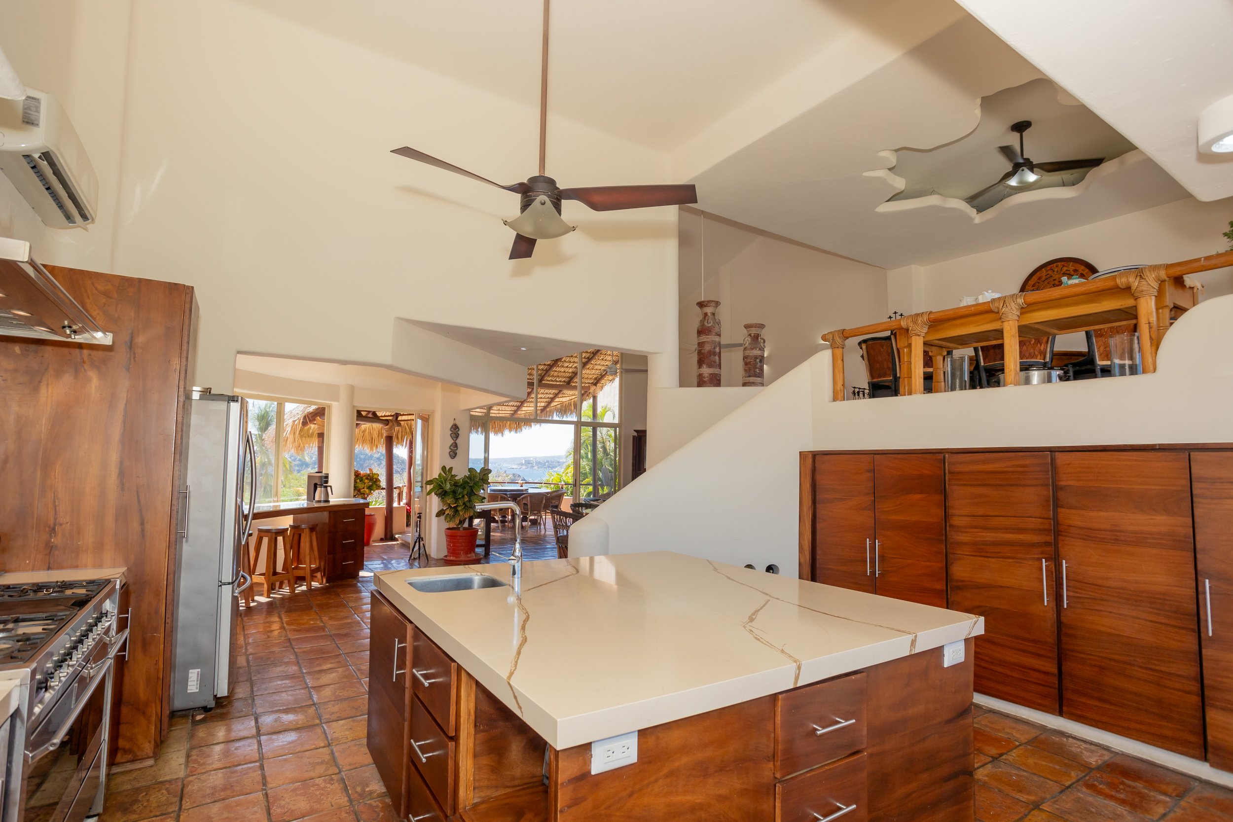 A spacious kitchen with a white marble island, wooden cabinets, and a view of an outdoor patio with thatched roofing and tropical scenery.