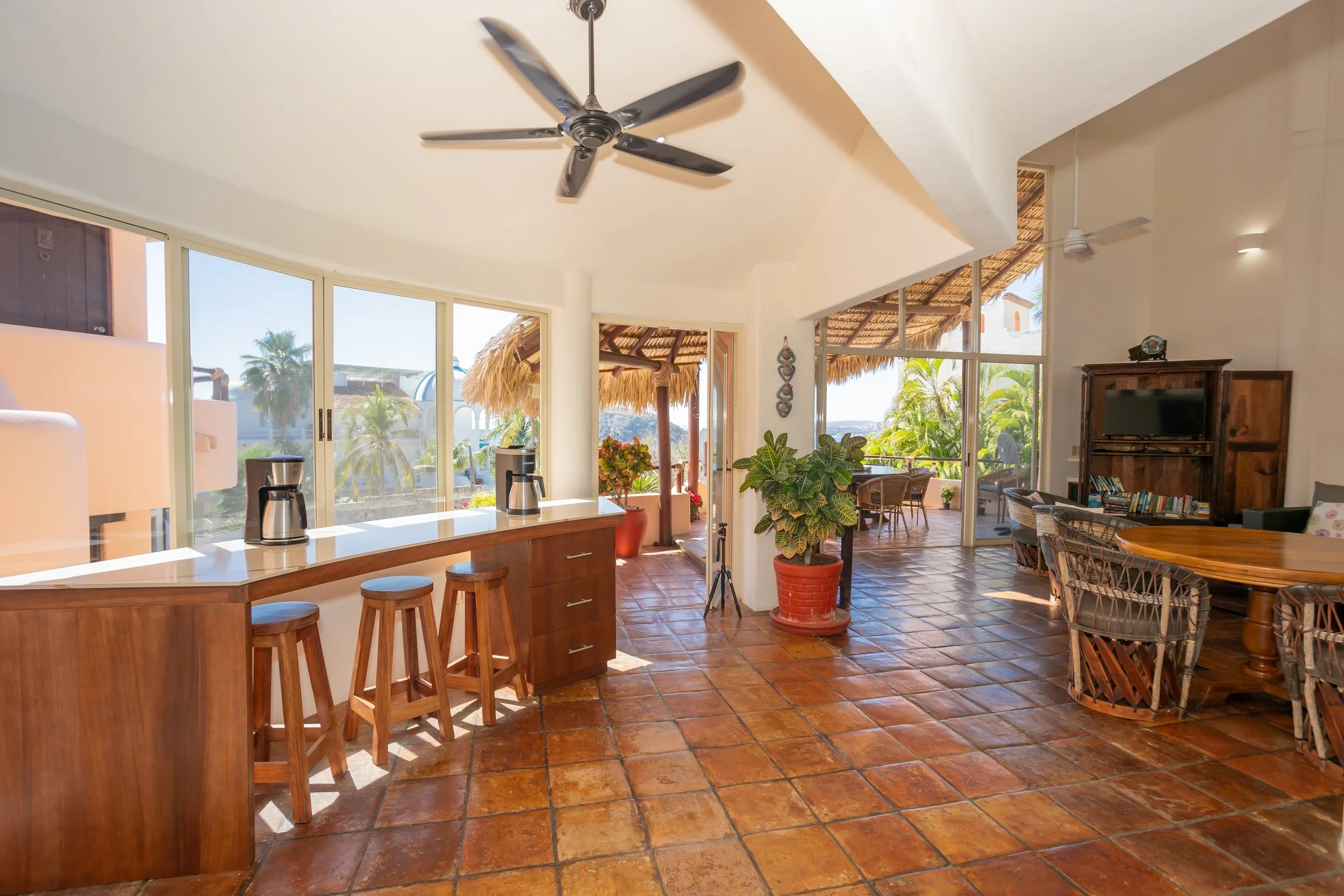 Bright living space with large windows, ceiling fans, potted plants, a kitchen island with stools, and a dining area with wicker chairs, during daytime with outdoors visible.