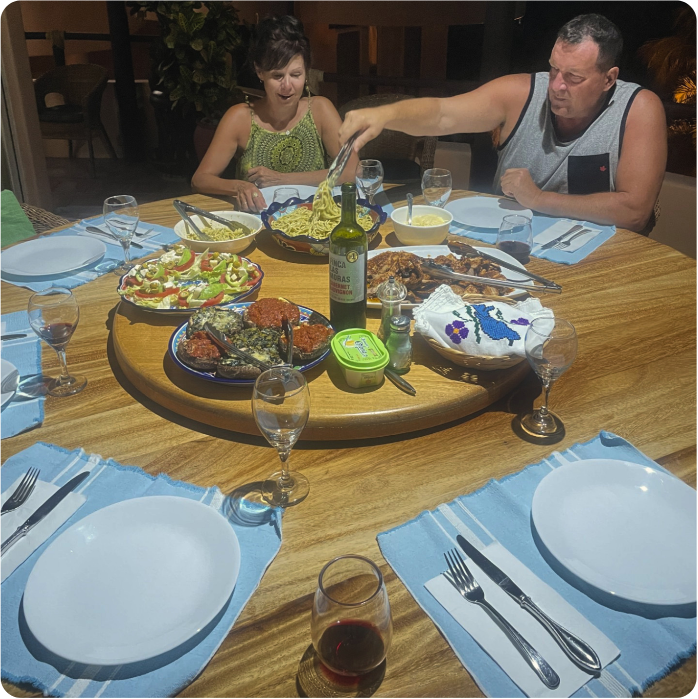 A dinner table with plates, glasses, utensils, and a variety of food including fish, vegetables, pasta, and wine. Two people are seated at the table, one man serving food and one woman reacting with surprise.