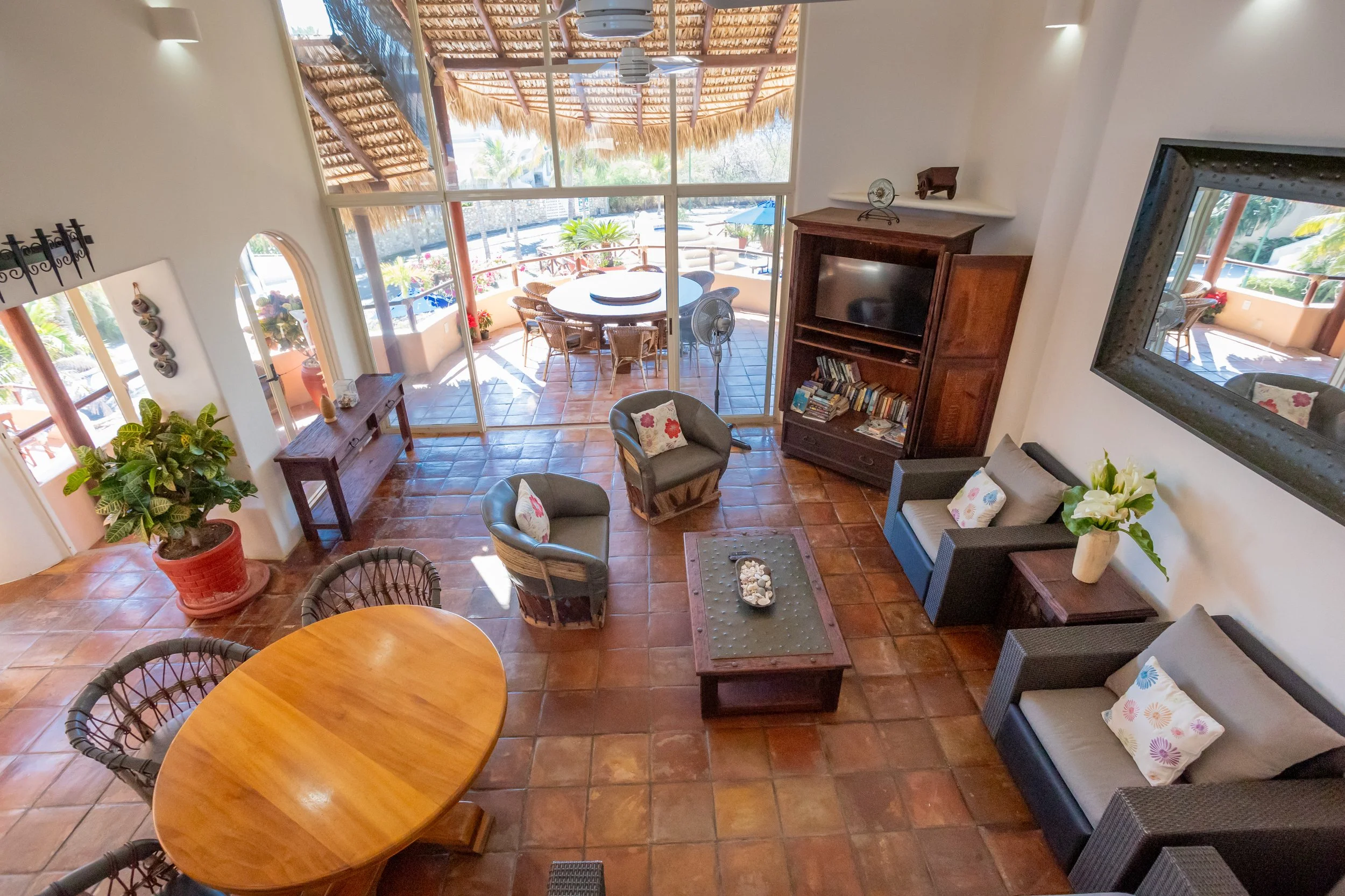 A cozy living room with terracotta tiled floor, a wooden round dining table with chairs, a grey couch with floral pillows, a small coffee table, and a large mirror on the wall. There is a tall bookshelf filled with books and a TV. Large windows and a