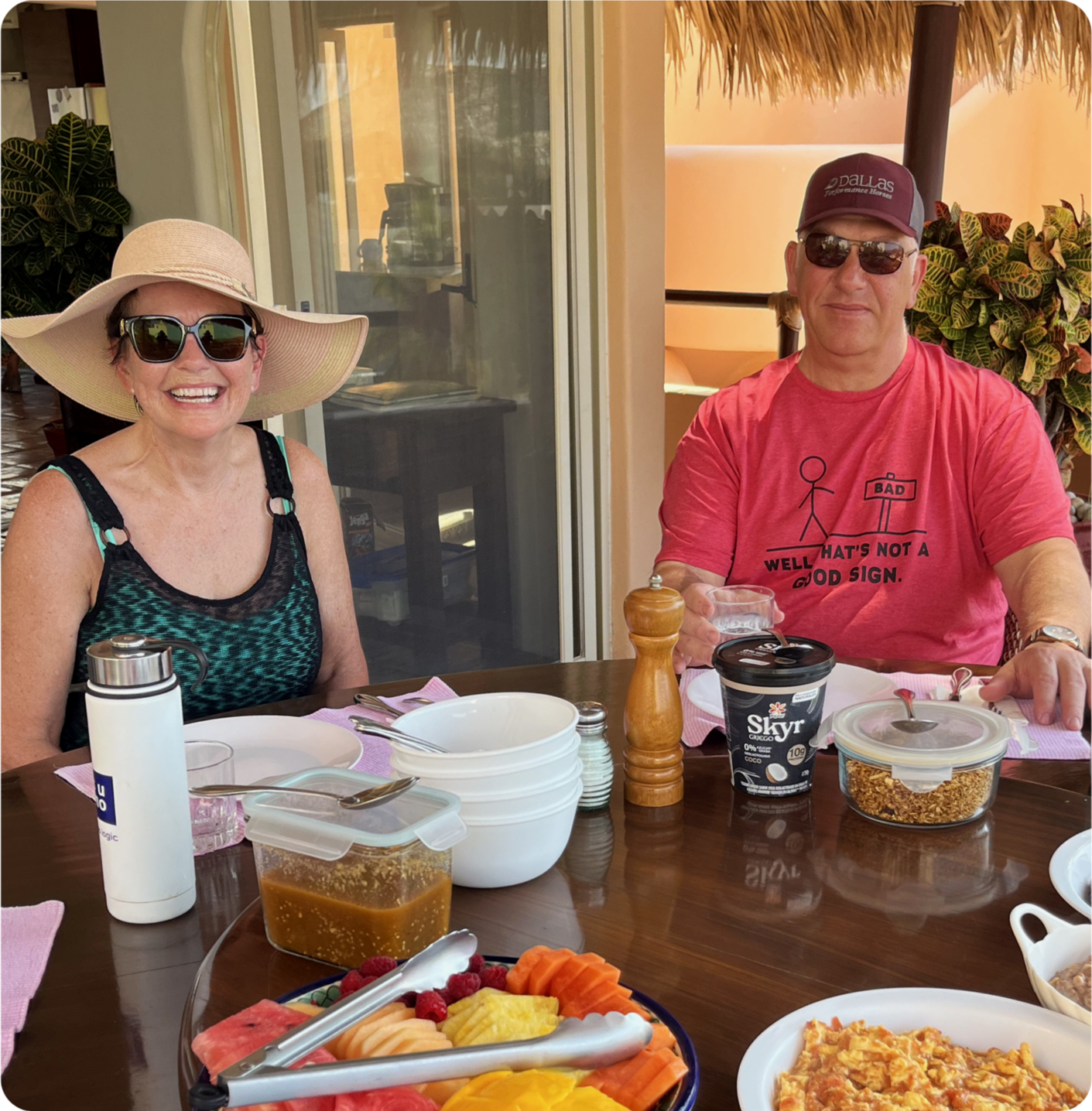 Two people sitting at a table outdoors, smiling. The woman on the left is wearing a large sunhat, sunglasses, and a black and green tank top. The man on the right is wearing a maroon cap, sunglasses, and a red T-shirt with a graphic and text. The table has various food items, including a fruit platter, a container of granola, a tub of Skyr yogurt, a pepper grinder, and a white thermos. The background shows a sliding glass door and some plants.