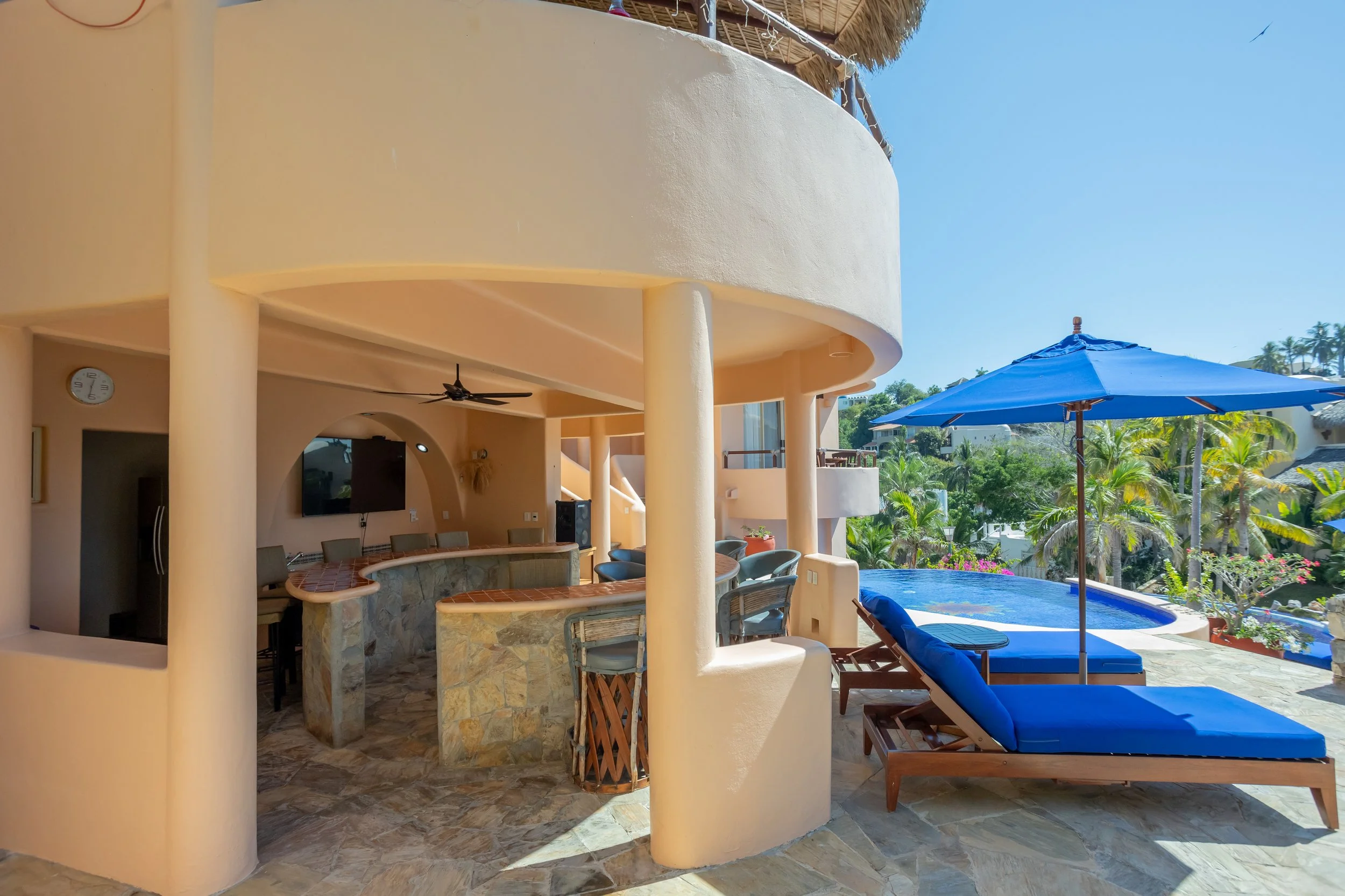 A backyard patio with a round bar, green chairs, a blue pool umbrella, and a circular pool, surrounded by tropical trees.