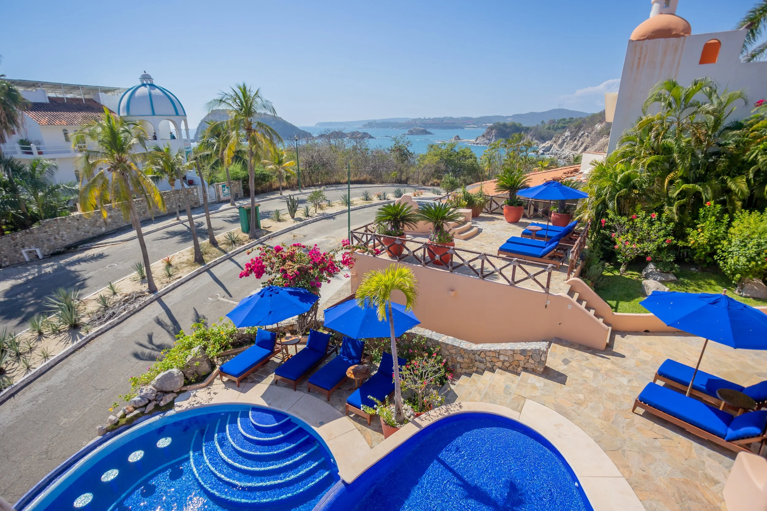 A scenic view of a coastal area with a terrace featuring blue lounge chairs and umbrellas, lush green plants, palm trees, and a small swimming pool, overlooking the ocean with rocky islands in the distance under a clear blue sky.