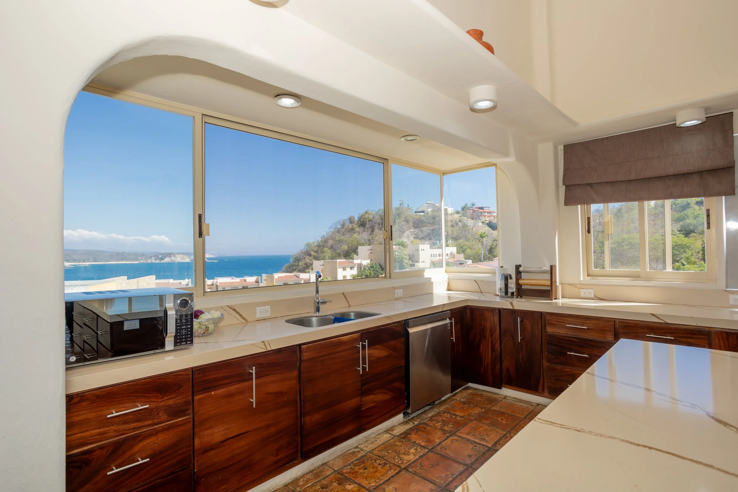Kitchen with large windows showing a view of hills, houses, and the ocean; wooden lower cabinets, white countertops, microwave, and kettle.
