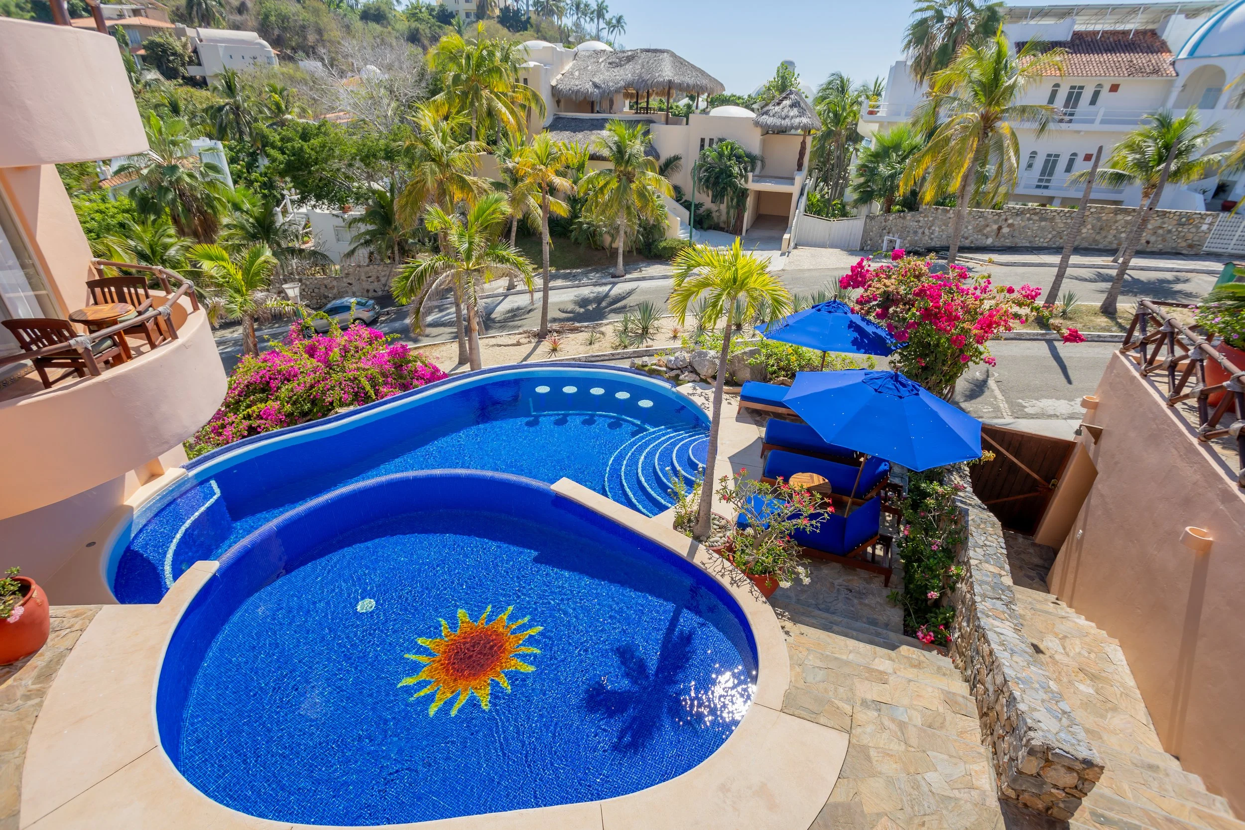 Balcony view overlooking a swimming pool with blue umbrellas, surrounded by pink bougainvillaea flowers, palm trees, and buildings in a tropical style.