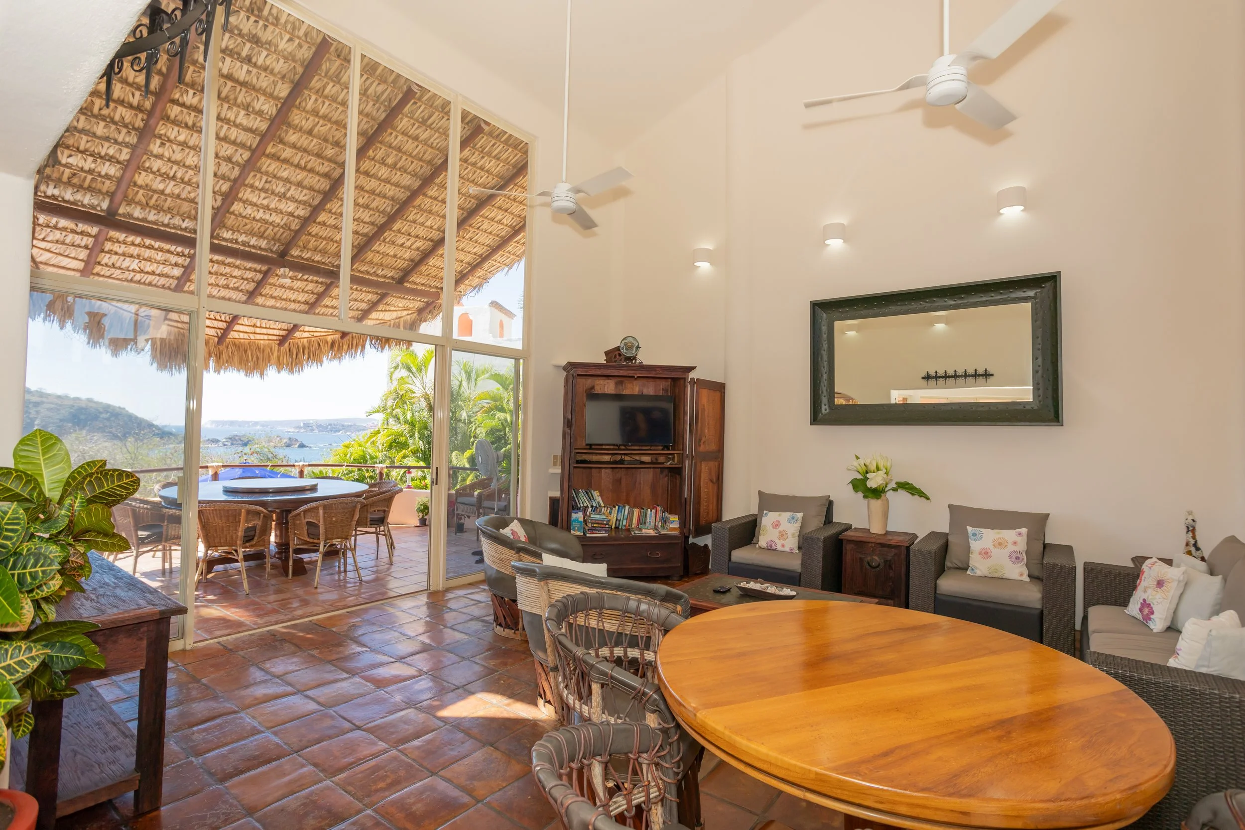 Living room with a large window showing a thatched roof patio outside, featuring a dining table, outdoor chairs, and greenery with a view of water and hills in the distance.