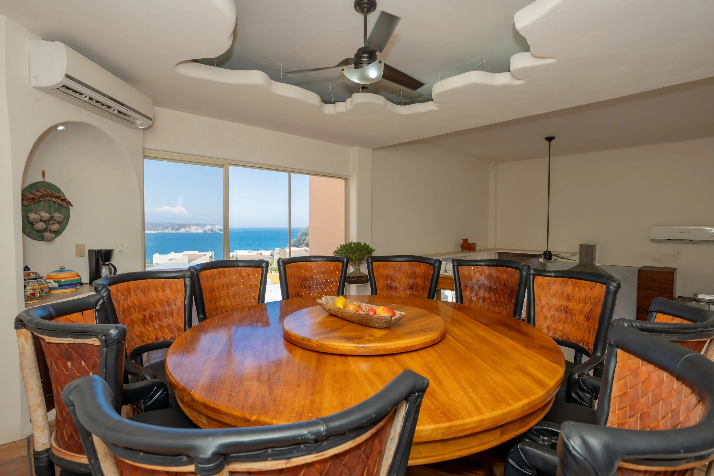 A dining room with a large round wooden table surrounded by black chairs, a bowl of fruit on the table, windows showing a view of the ocean and coastline, a ceiling fan, and decorative items on the wall and counter.