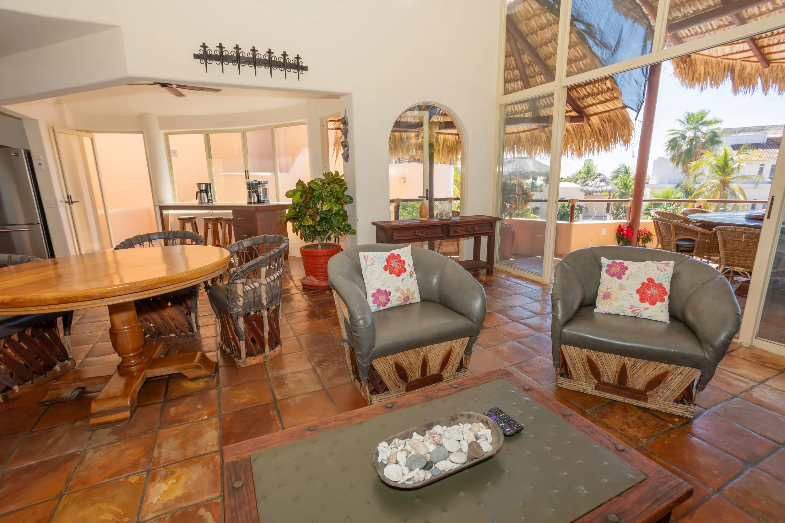 Living room with large windows and a balcony, featuring wicker chairs with floral cushions, a wooden dining table with chairs, a potted plant, and tropical scenery outside.