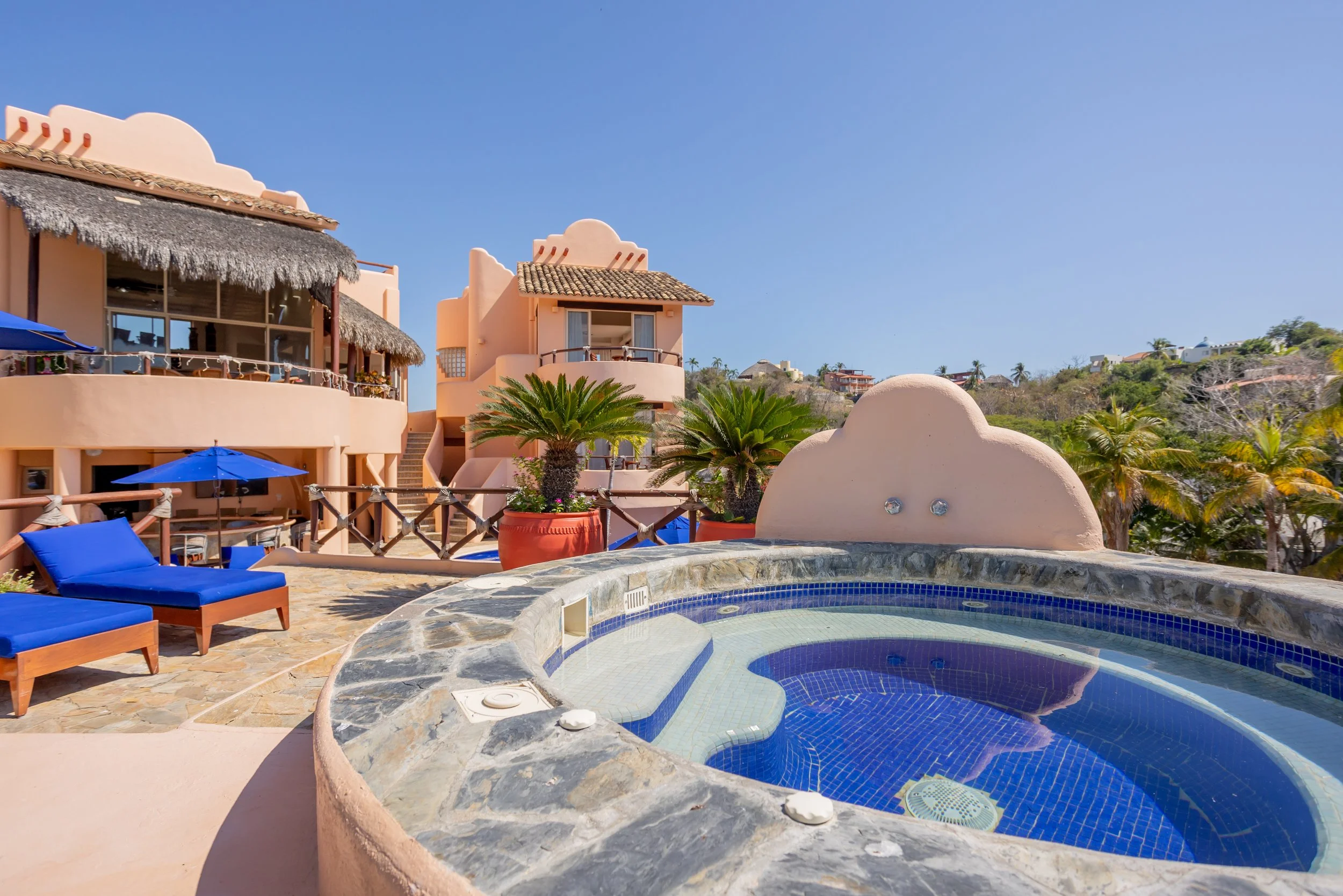 A rooftop view of a hotel or resort with a small swimming pool, blue lounge chairs, palm trees, and pink buildings with thatched and tiled roofs under a clear blue sky.