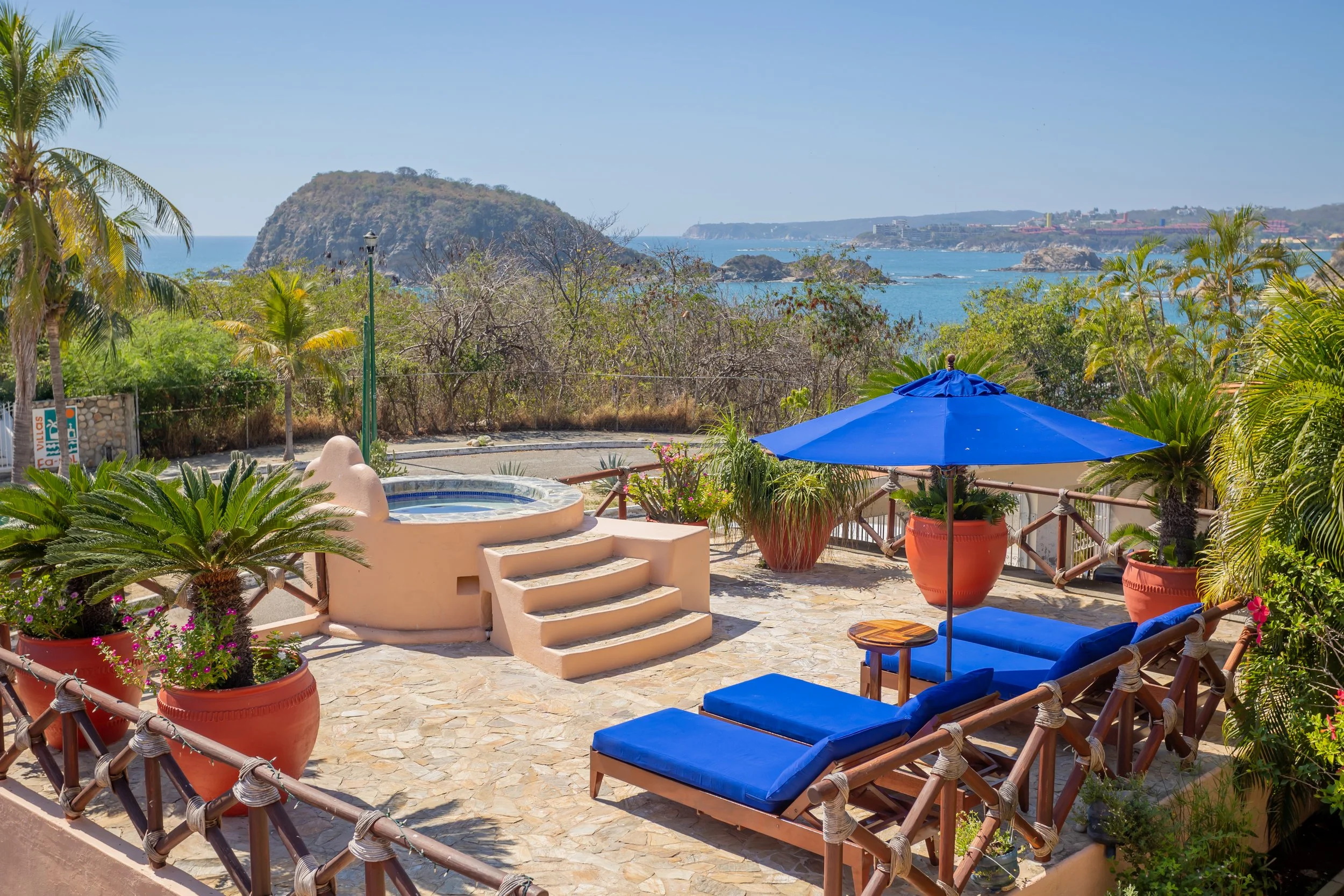 A scenic ocean view with lush greenery, a rocky island in the distance, and a balcony with blue cushioned lounge chairs, potted plants, a blue umbrella, and a hot tub.