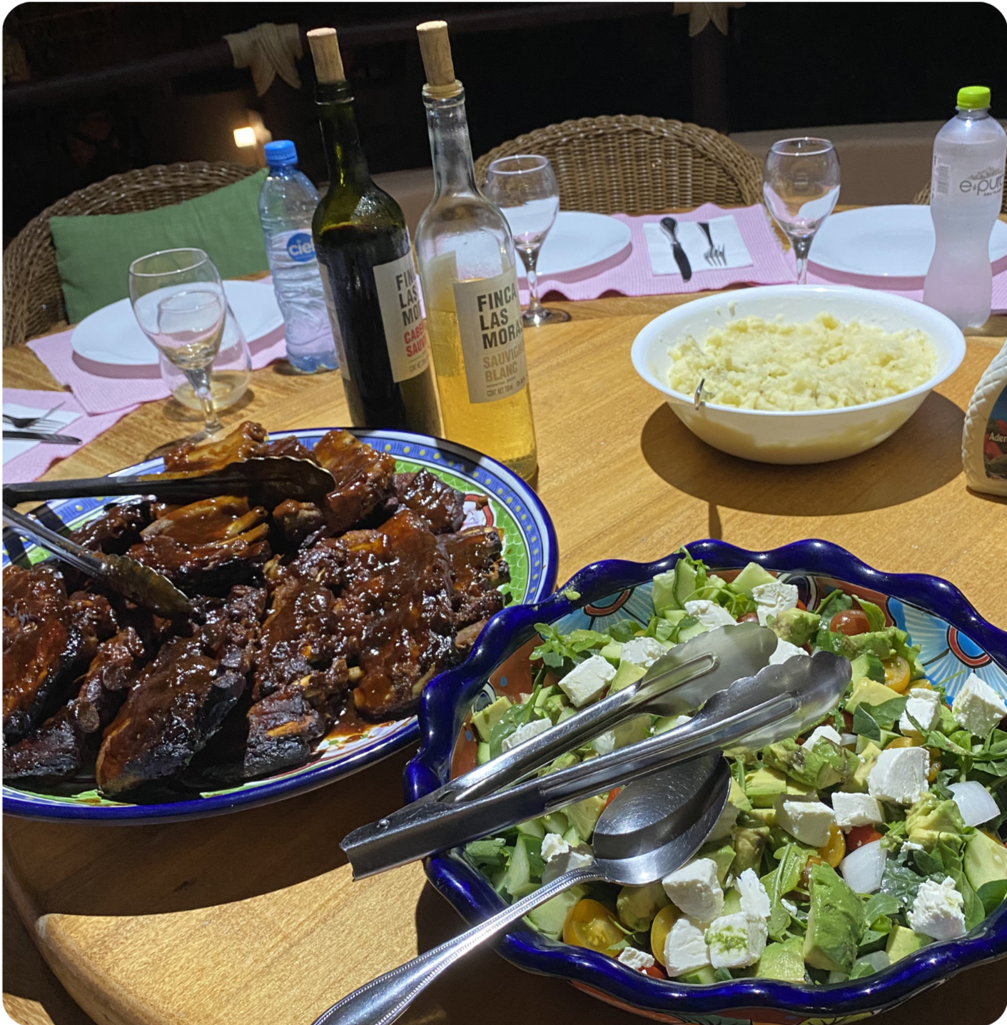 A table set with various dishes, including a bowl of mashed potatoes, a salad with cheese, cherry tomatoes, lettuce, and other greens, and a large dish of glazed meat ribs or chicken. There are bottles of wine, water, and condiments on the table, with plates, glasses, and utensils prepared for a meal.