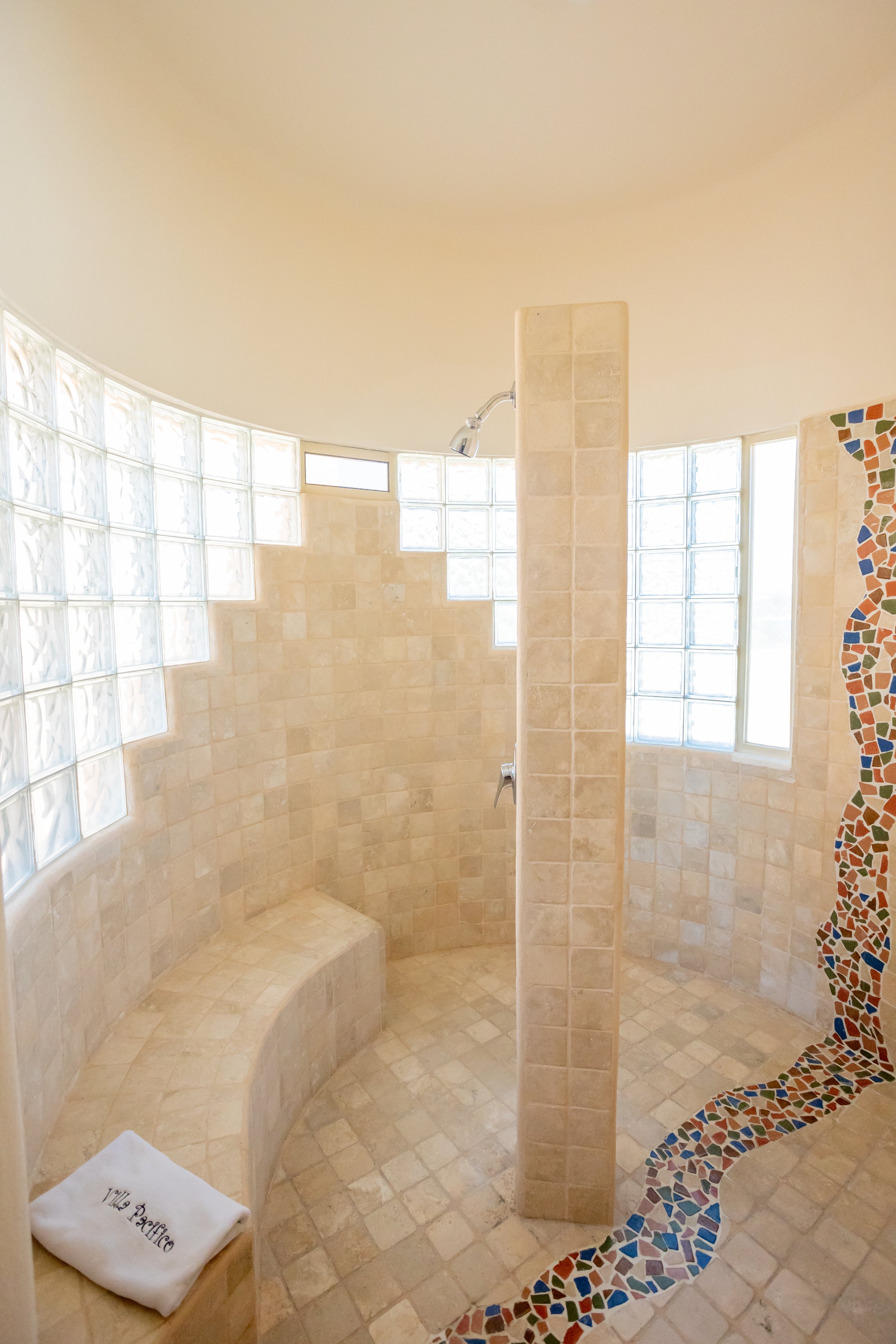 A shower and seating area with beige tiled walls and curved bench seating, light-colored windows, and a mosaic tile design on the curved wall.