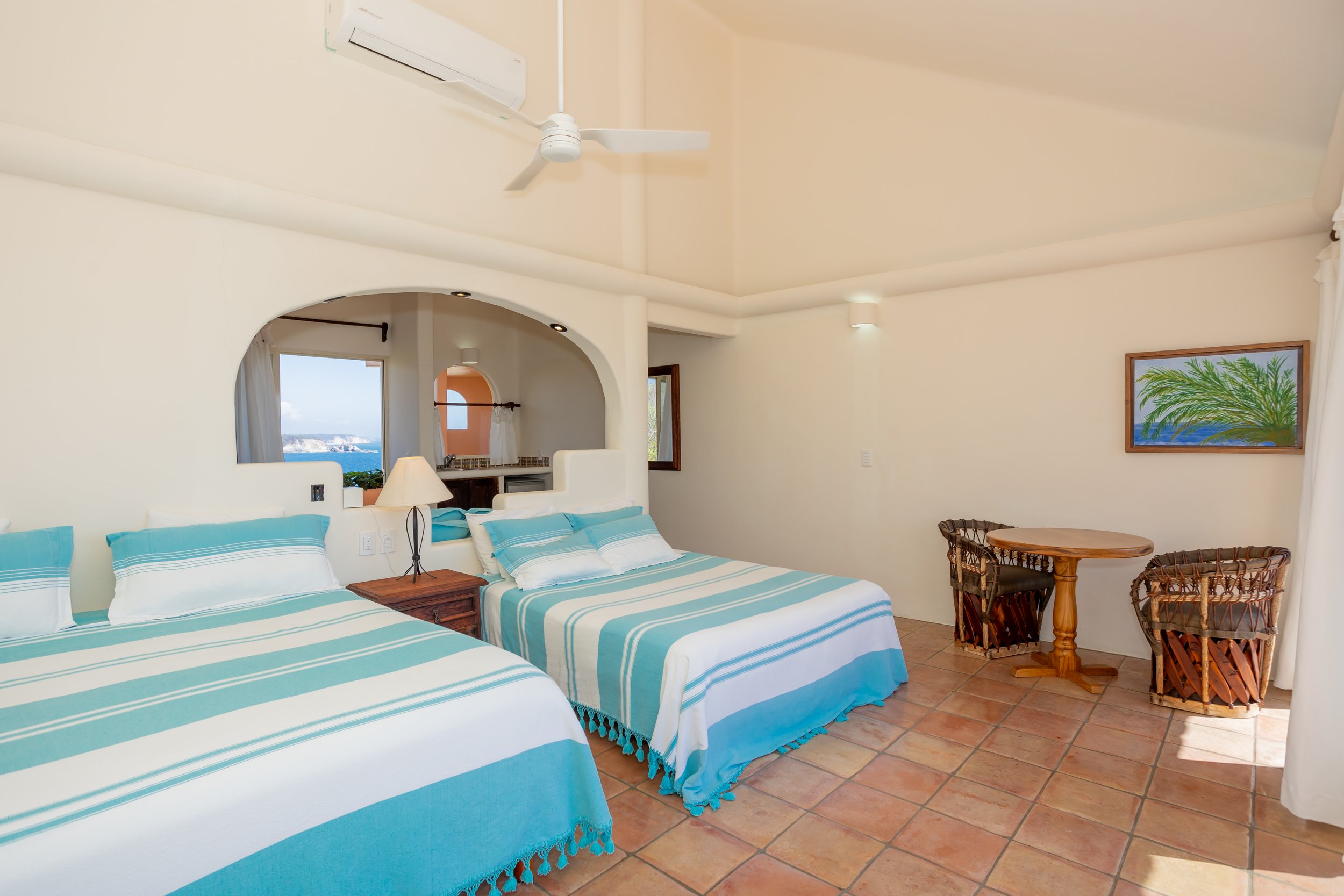A hotel room with two single beds with blue and white striped bedspreads, a nightstand and a lamp between the beds, a small round table with two wicker chairs near the wall, and a painting of a palm tree with a blue ocean background on the wall. There are arched openings, one with a view of the ocean and sky outside, and a ceiling fan in the room.