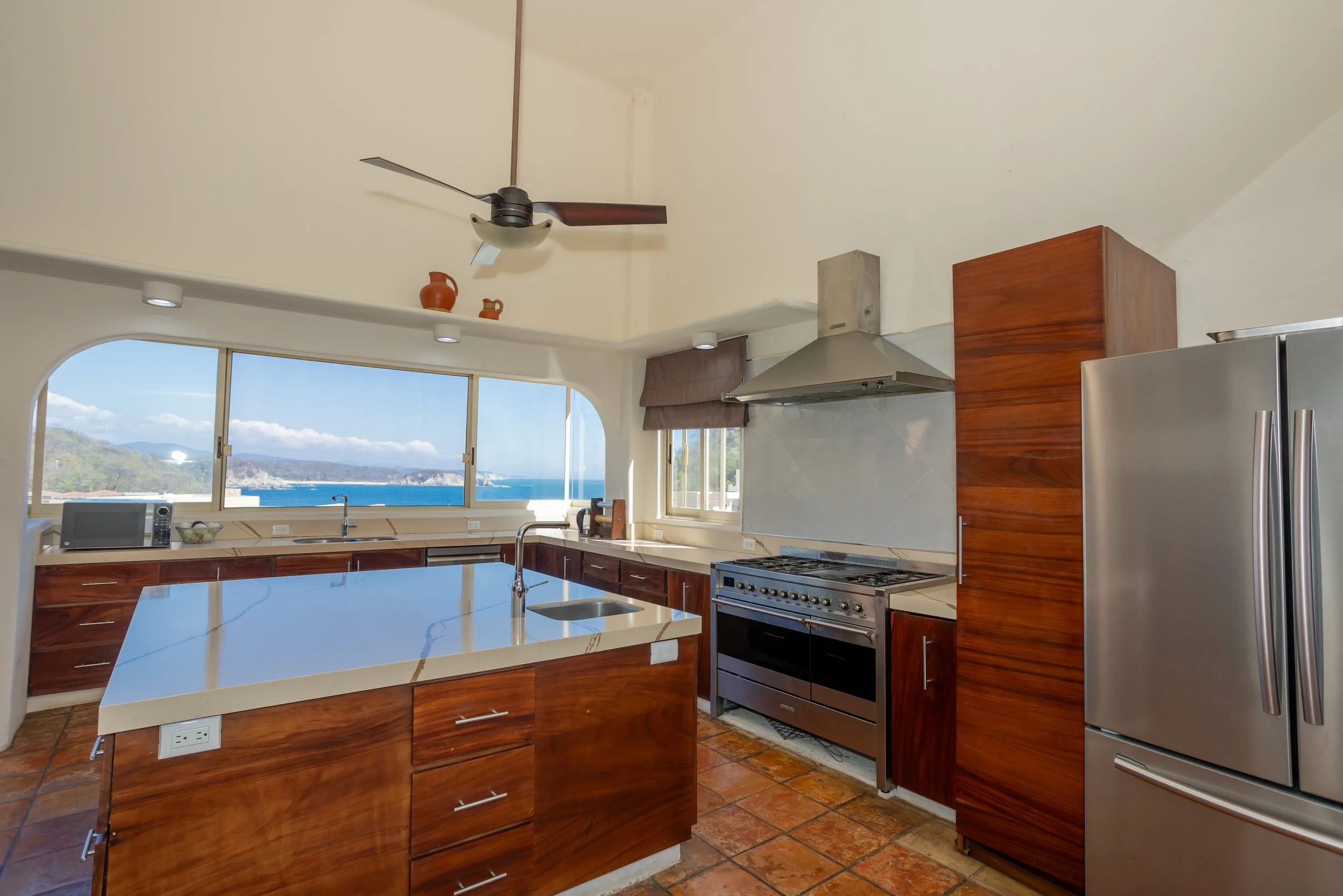 Modern kitchen with large window, wooden cabinetry, stainless steel appliances, and ocean view.
