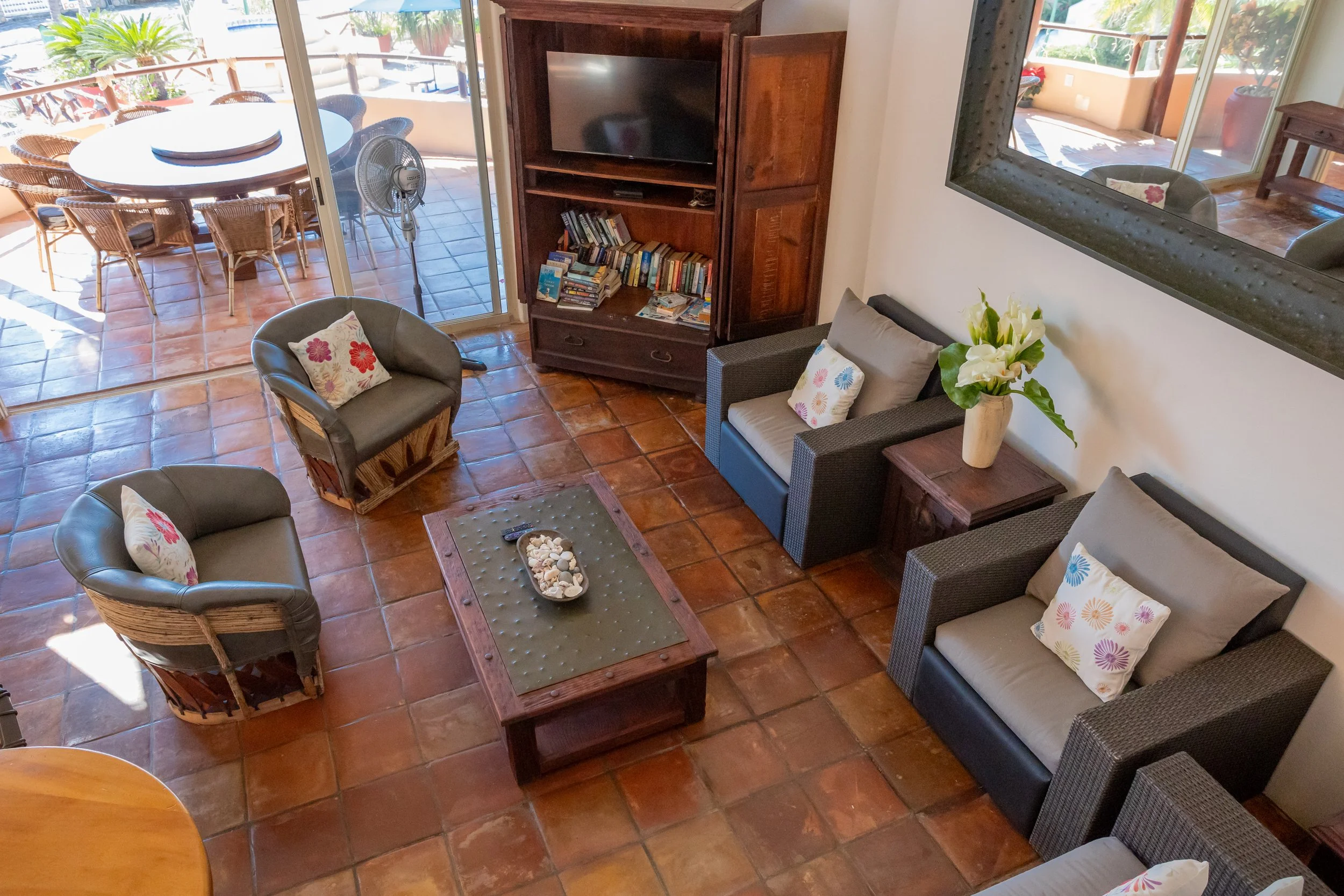 Living room with terracotta tile flooring, beige and black seating with floral cushions, a wooden coffee table with seashells, a large mirror, a wooden entertainment center with a TV and books, and sliding glass doors leading to an outdoor patio with