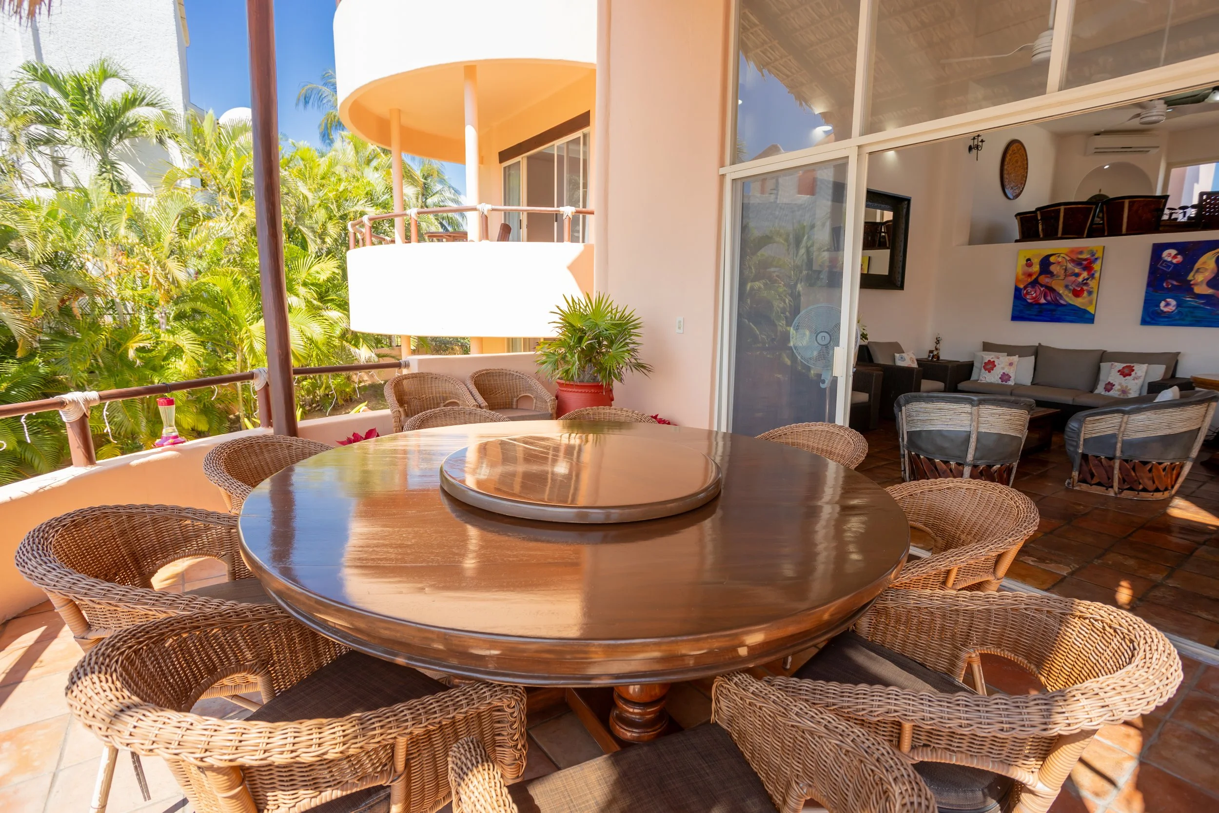 A view of a balcony with a round wooden table and wicker chairs, overlooking lush green tropical plants with a building in the background. Inside, a living room with artwork, a couch, and a sliding glass door allowing natural light.