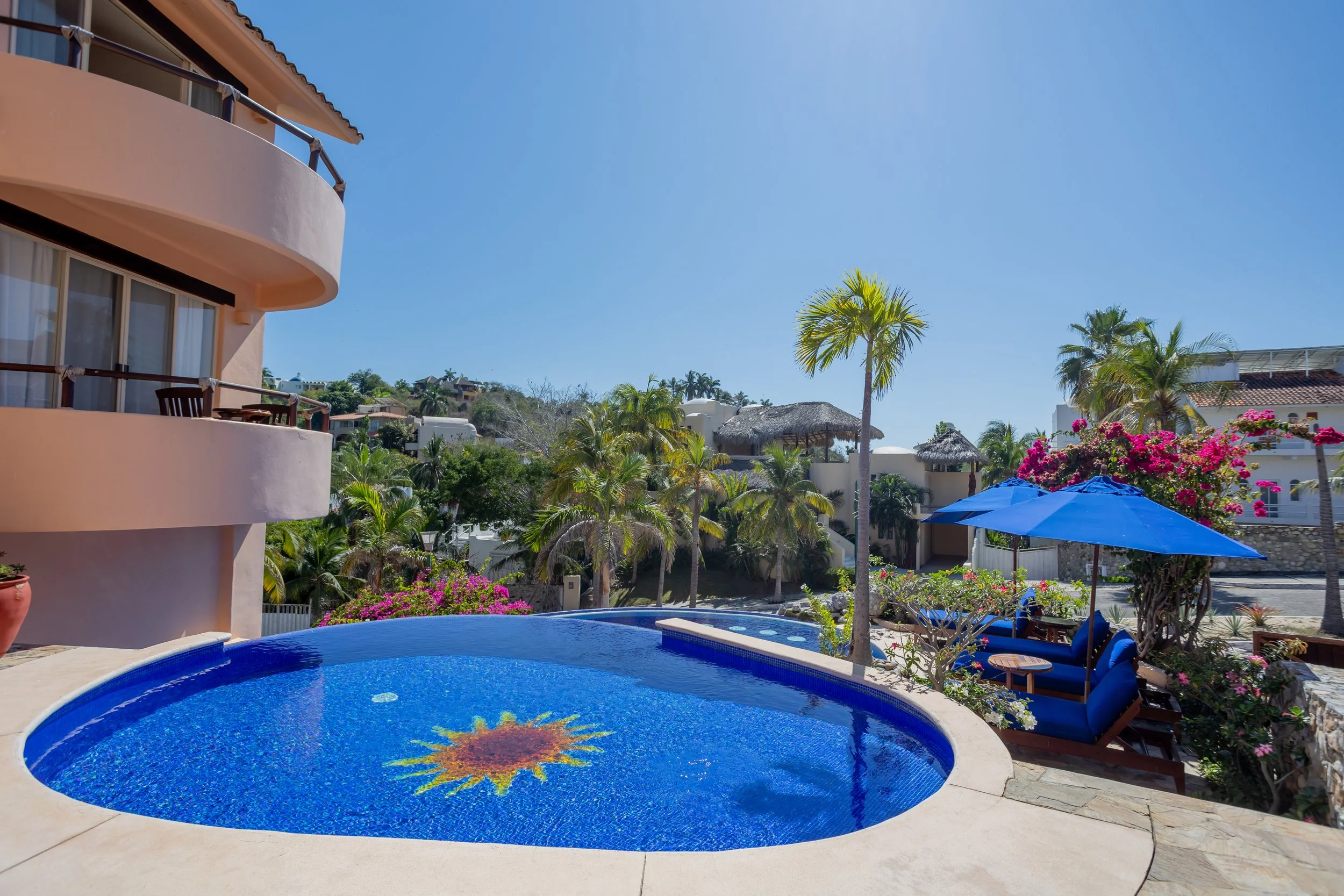 A swimming pool with a mosaic sun design at the bottom, surrounded by colorful flowers, blue umbrellas, and lounge chairs, with tropical plants and buildings in the background under a clear blue sky.